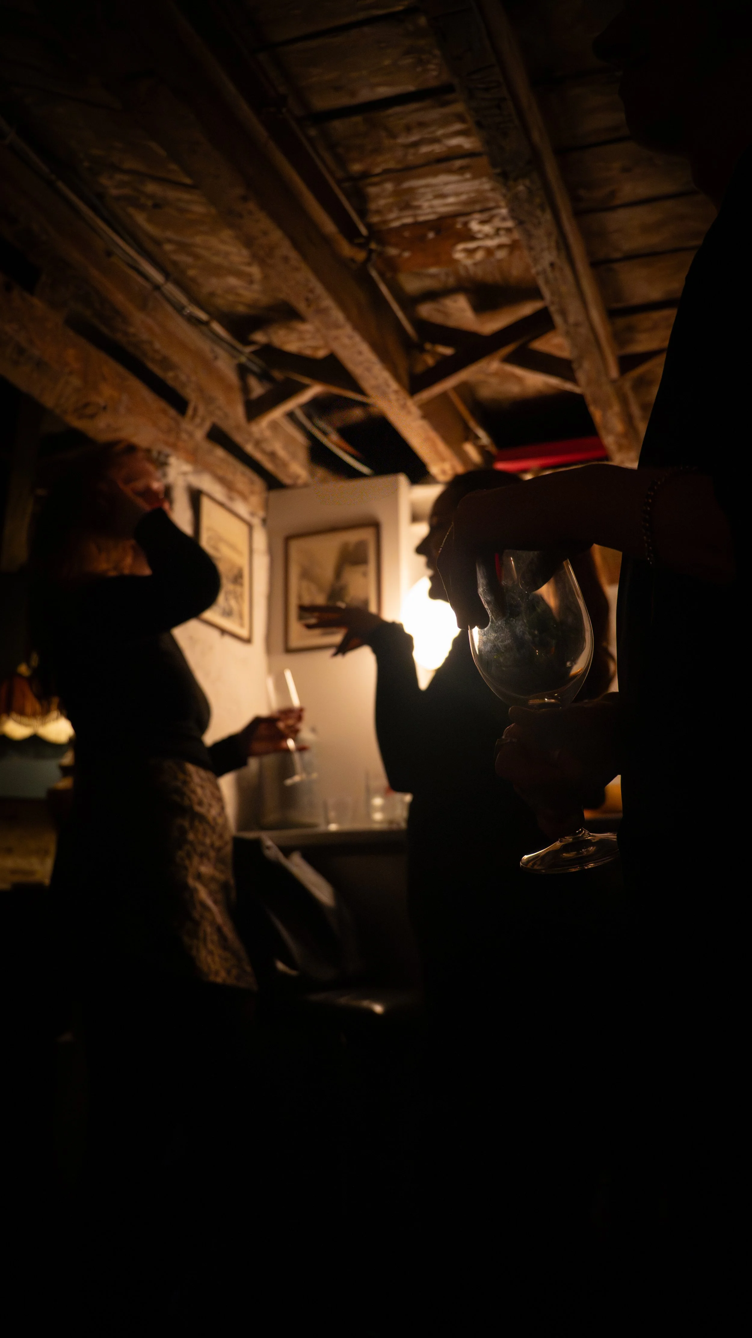 People socializing in a dimly lit, rustic setting with wooden beams and framed pictures on the wall, holding drinks.