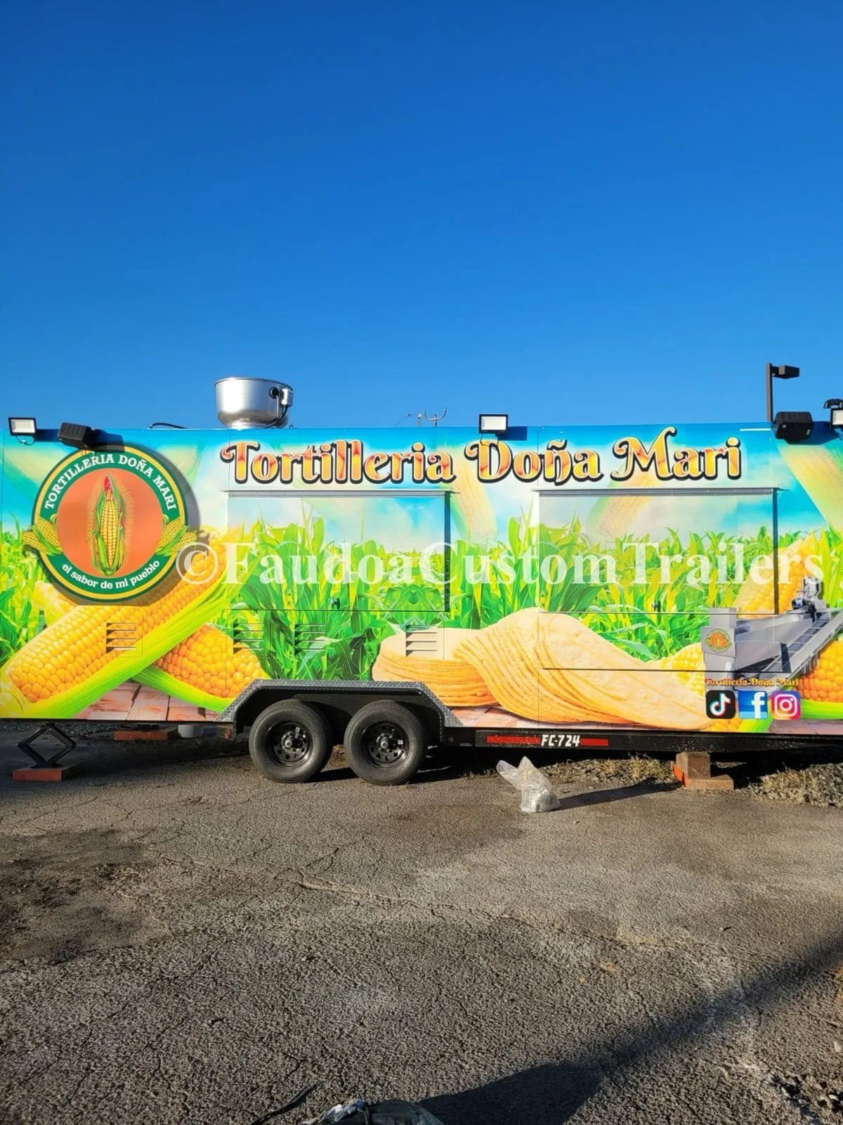 A colorful food truck with a graphic of corn and chips, and the text "Tortilleria Dóna Mari" on it, parked on a paved surface under a clear blue sky.