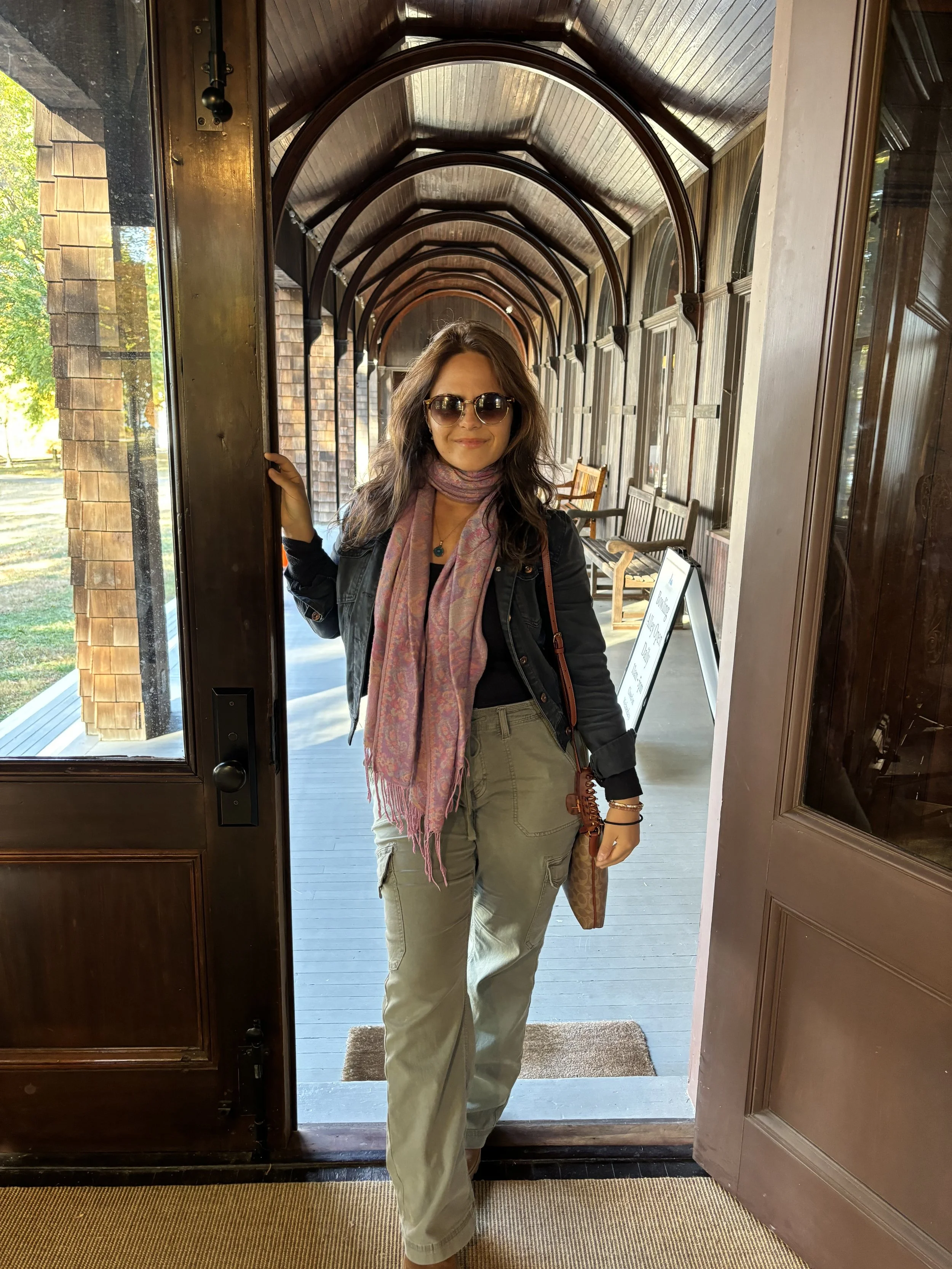 Woman standing in doorway of a wooden building, wearing sunglasses, a black jacket, khaki cargo pants, and a pink scarf.