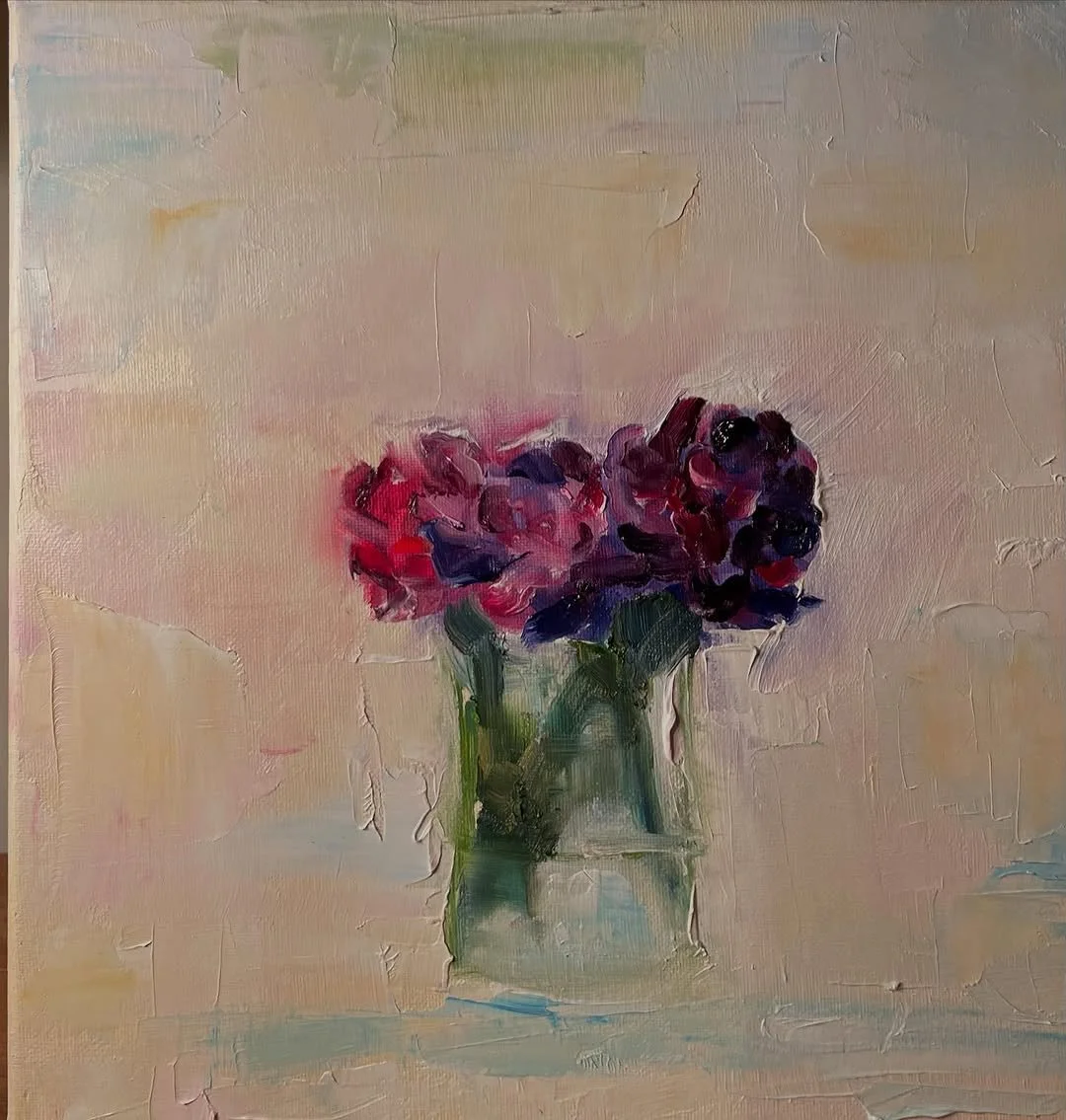 A still life painting of a bouquet of purple and pink flowers in a glass vase on a light-colored surface with a soft, pastel background.