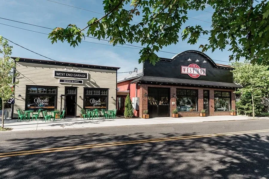 A street view featuring two establishments: 'West End Garage Bakery & Cafe' on the left with outdoor seating, and 'West End' on the right with large windows and brick accents, partly shaded by tree branches.