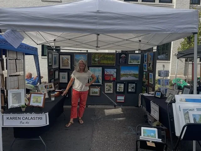A woman standing in front of an art booth displaying framed paintings at an outdoor art fair.