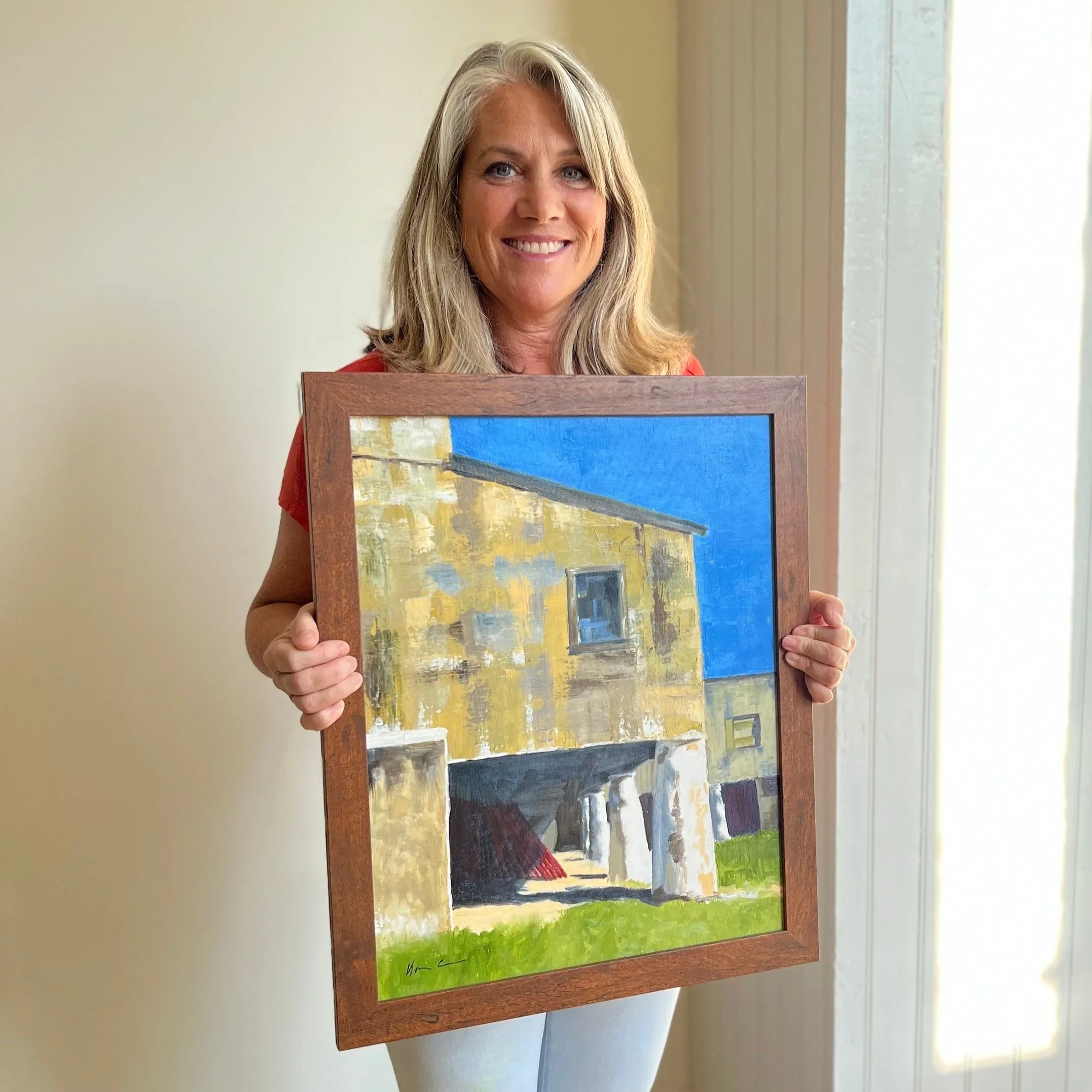 A smiling woman with shoulder-length blond hair holding a framed painting of a building under a bright blue sky.