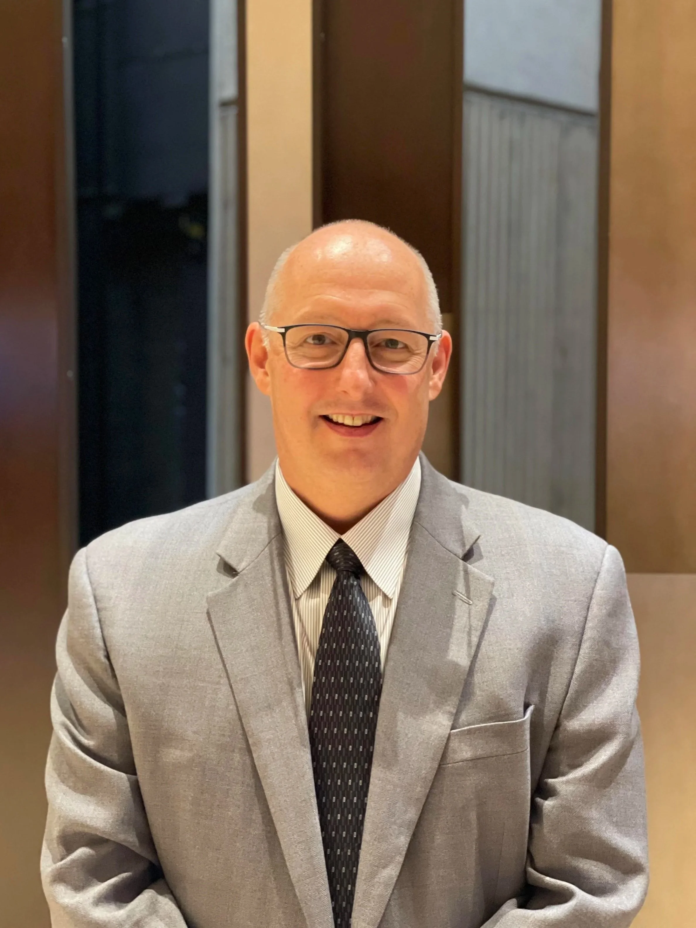 A man wearing glasses, a light grey suit, a white shirt, and a dark tie, smiling, standing indoors with wood-paneled walls in the background.