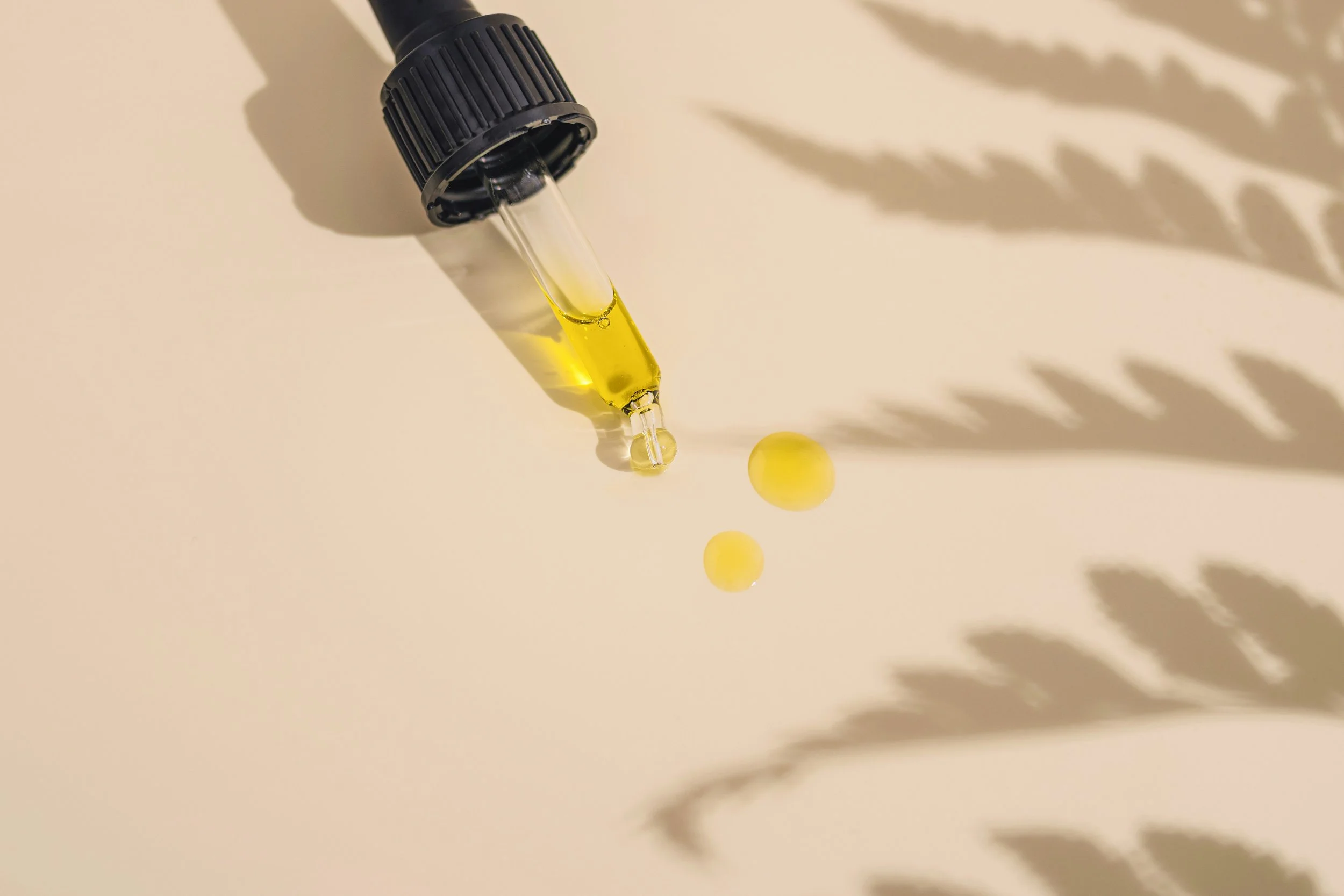 A dropper with yellow liquid above two yellow spherical capsules on a beige surface, with shadows of leaves in the background.