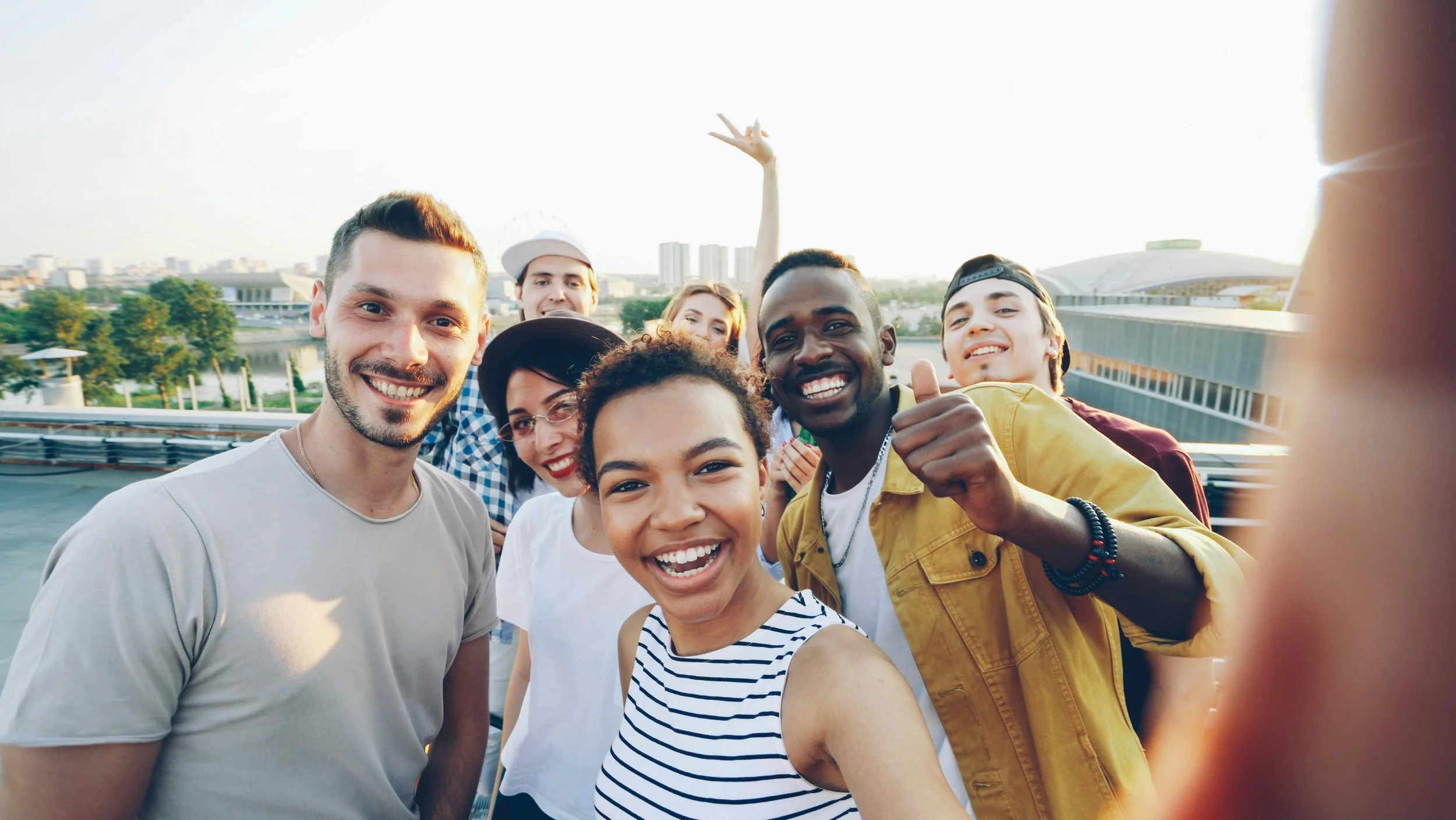 A diverse group of nine young people taking a selfie outdoors on a sunny day, smiling and having fun.
