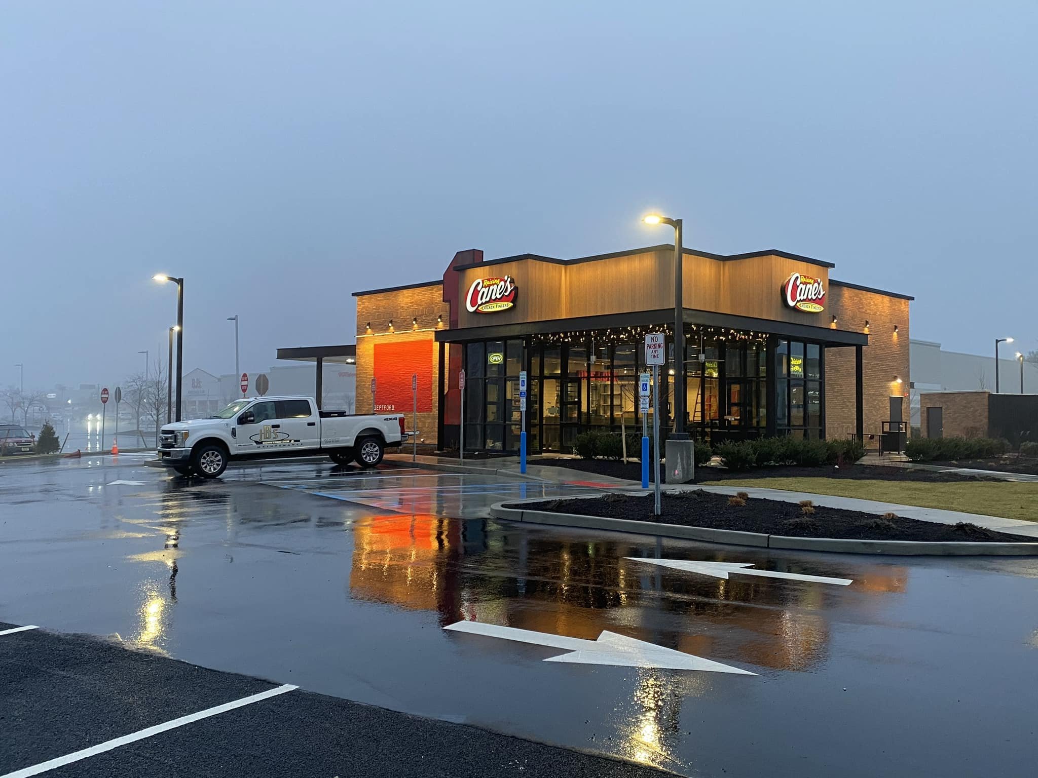 Exterior of a Raising Cane's restaurant in rainy weather, with a white pickup truck parked outside, wet pavement reflecting the building's lights, and a foggy background.