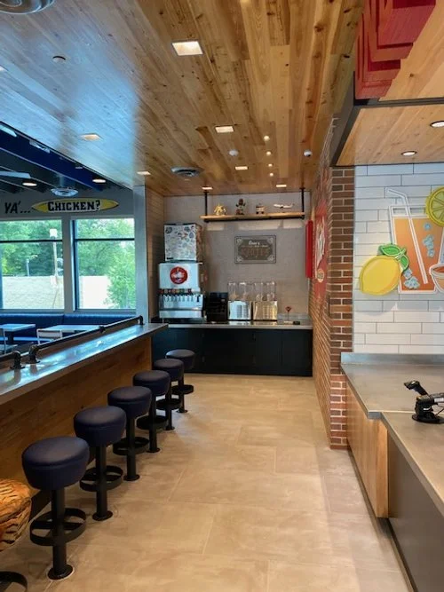Interior of a casual restaurant with a wood-paneled ceiling, seating area with stools, a beverage station with glasses and a fridge, and colorful wall art including fruit illustrations.