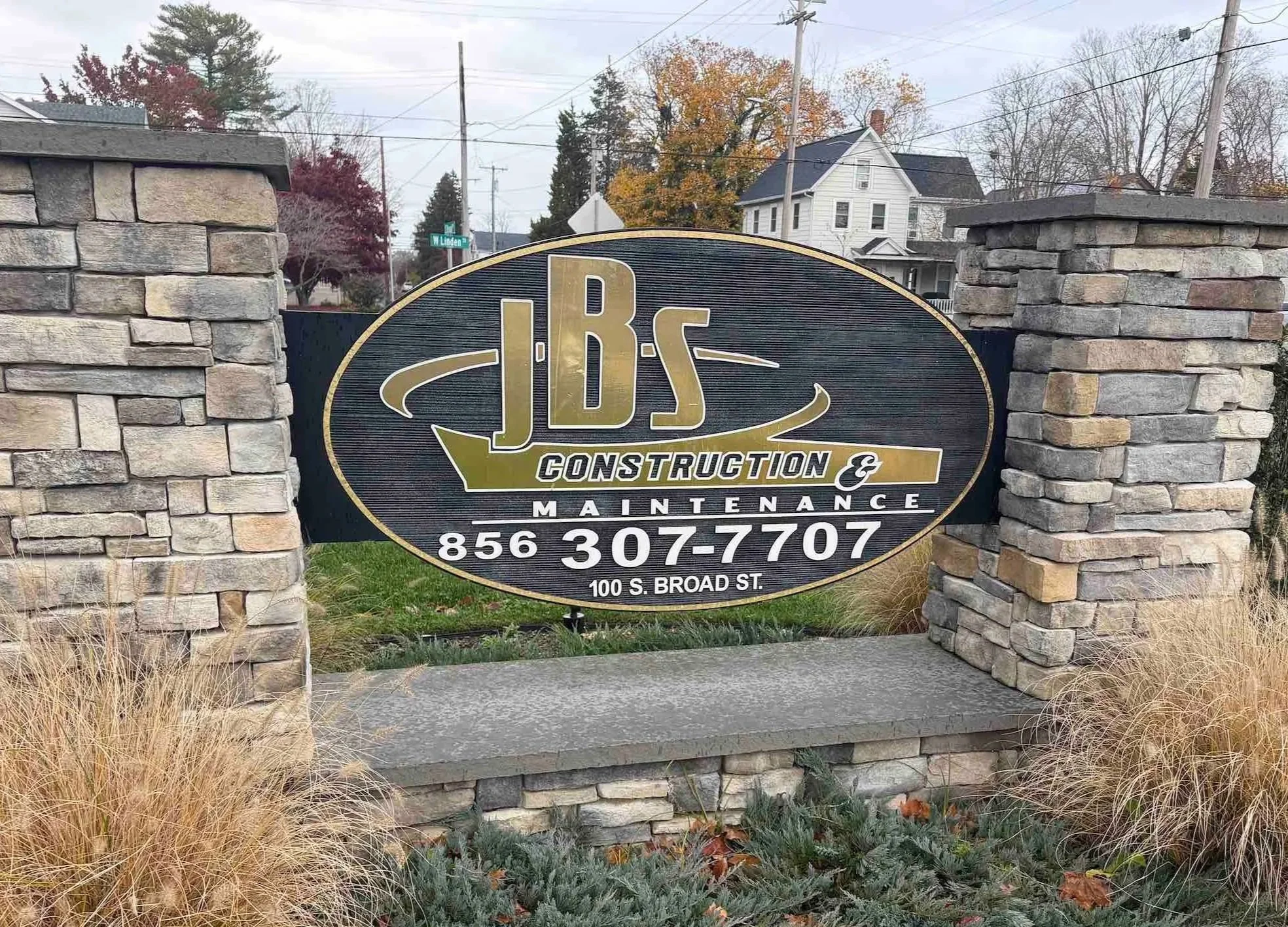 Sign for JBS Construction & Maintenance mounted between two stone pillars, with a street and houses visible in the background.