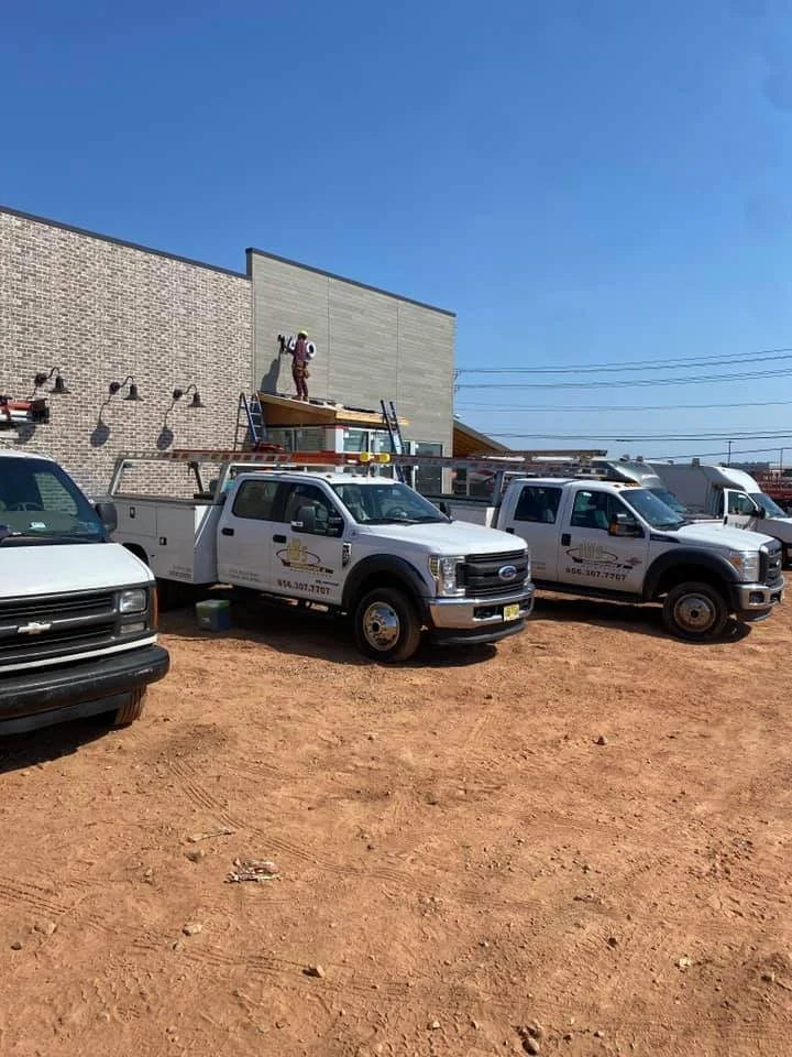 Worker on a ladder painting or repairing the exterior wall of a commercial building. Several utility trucks are parked nearby on a dirt lot.