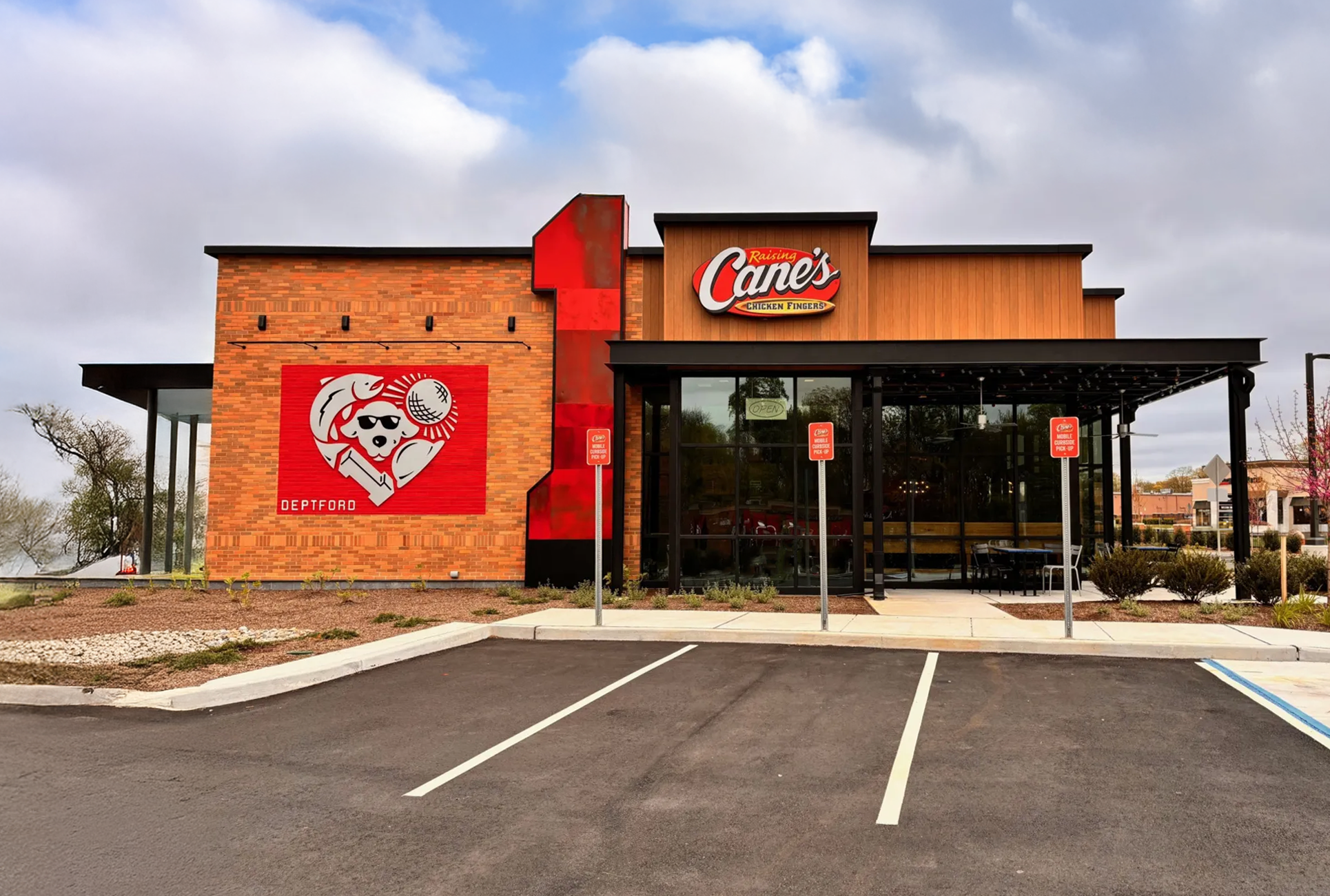 Exterior of Raising Cane's restaurant with brick and wood facade, large Cane's logo, and parking spaces in front.