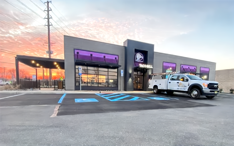 Taco Bell restaurant with purple accents, parked white service van in front, and apparent sunset in the background.
