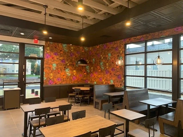 Interior of a modern cafe or restaurant with wooden tables and chairs, colorful abstract wallpaper, pendant lighting, large windows, and a door with an exit sign.