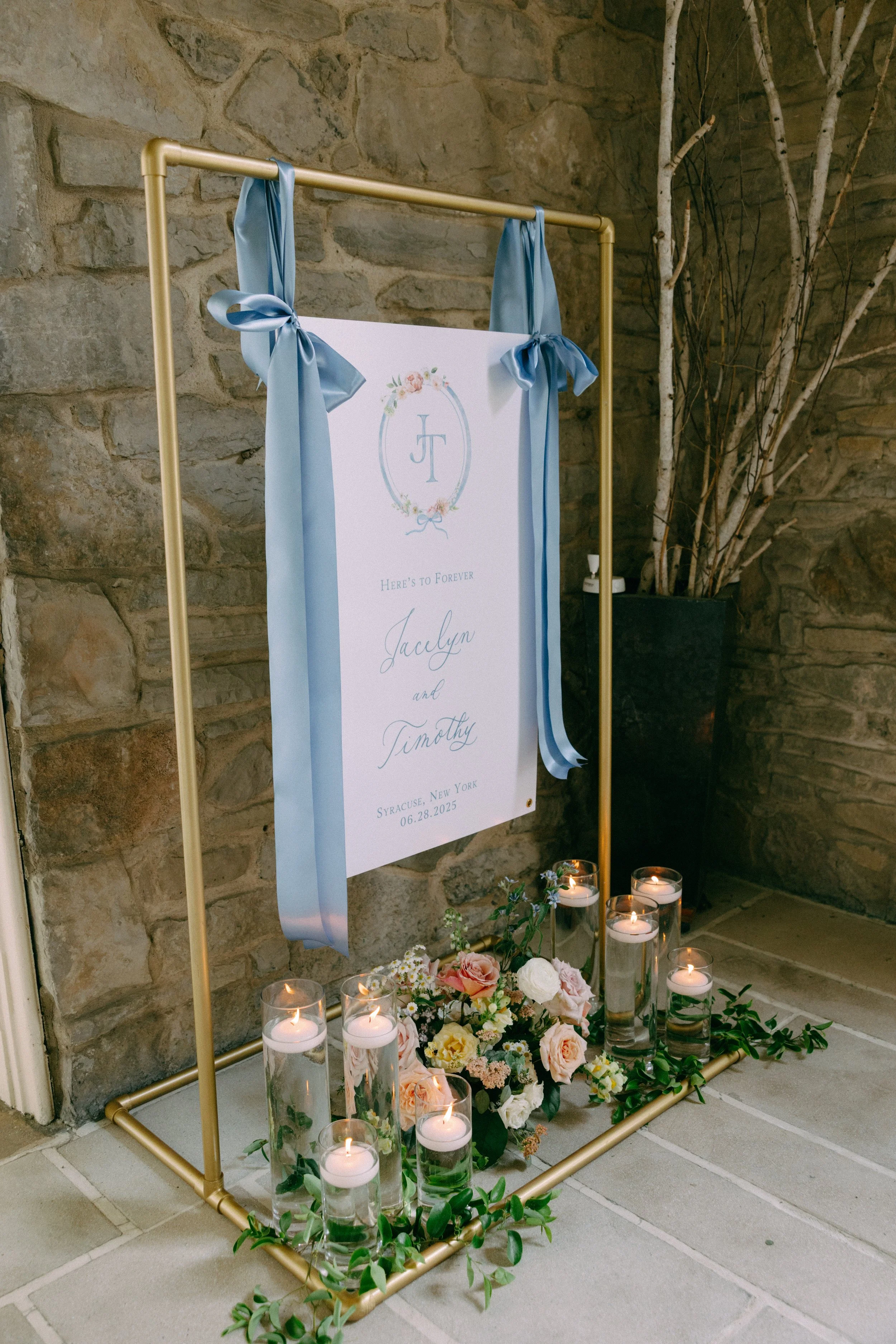 Wedding sign with blue ribbons, surrounded by candles and flowers, against a stone wall.