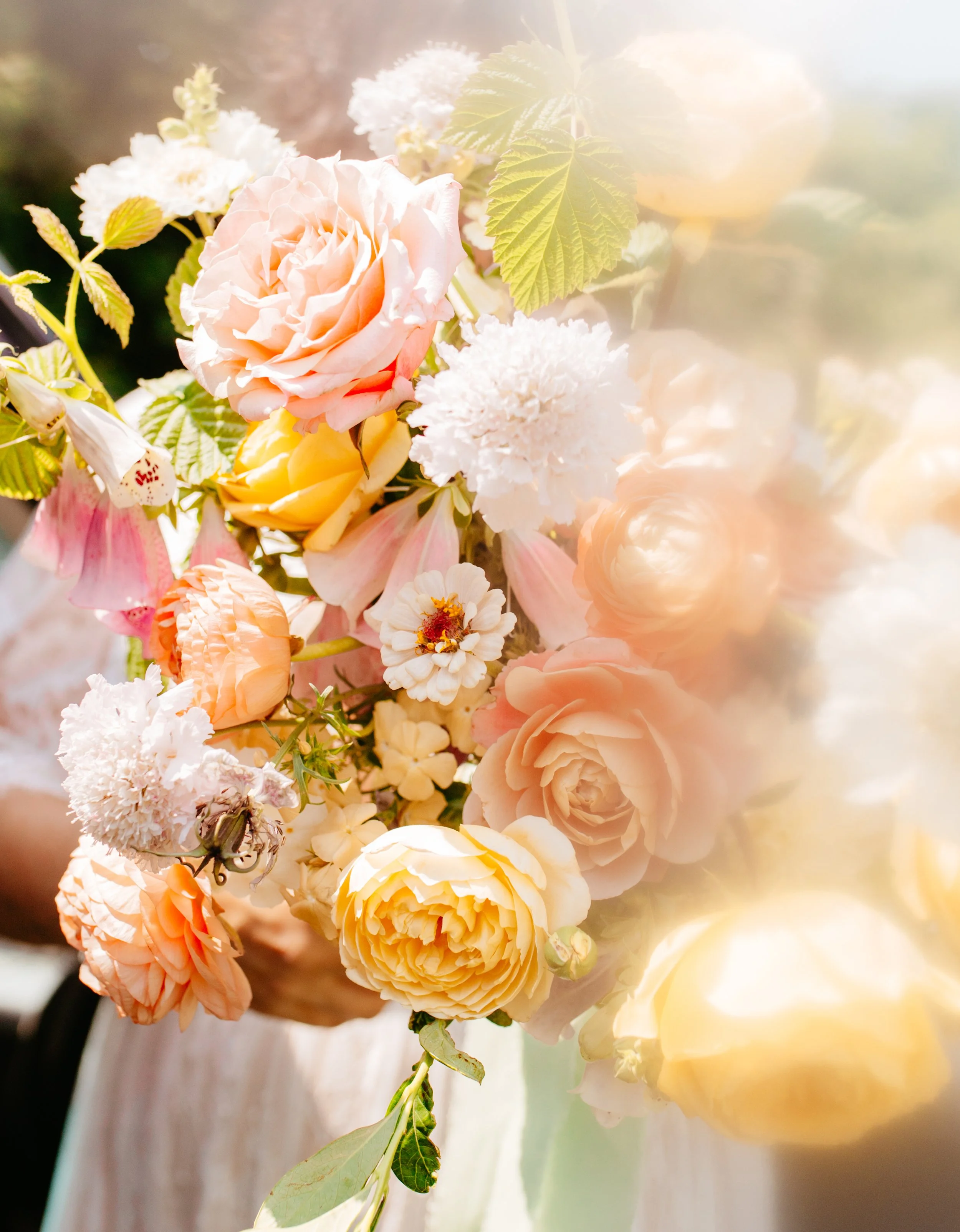 A bouquet of pastel-colored flowers including roses, peonies, and other blossoms, with green leaves, set against a soft, bright background.