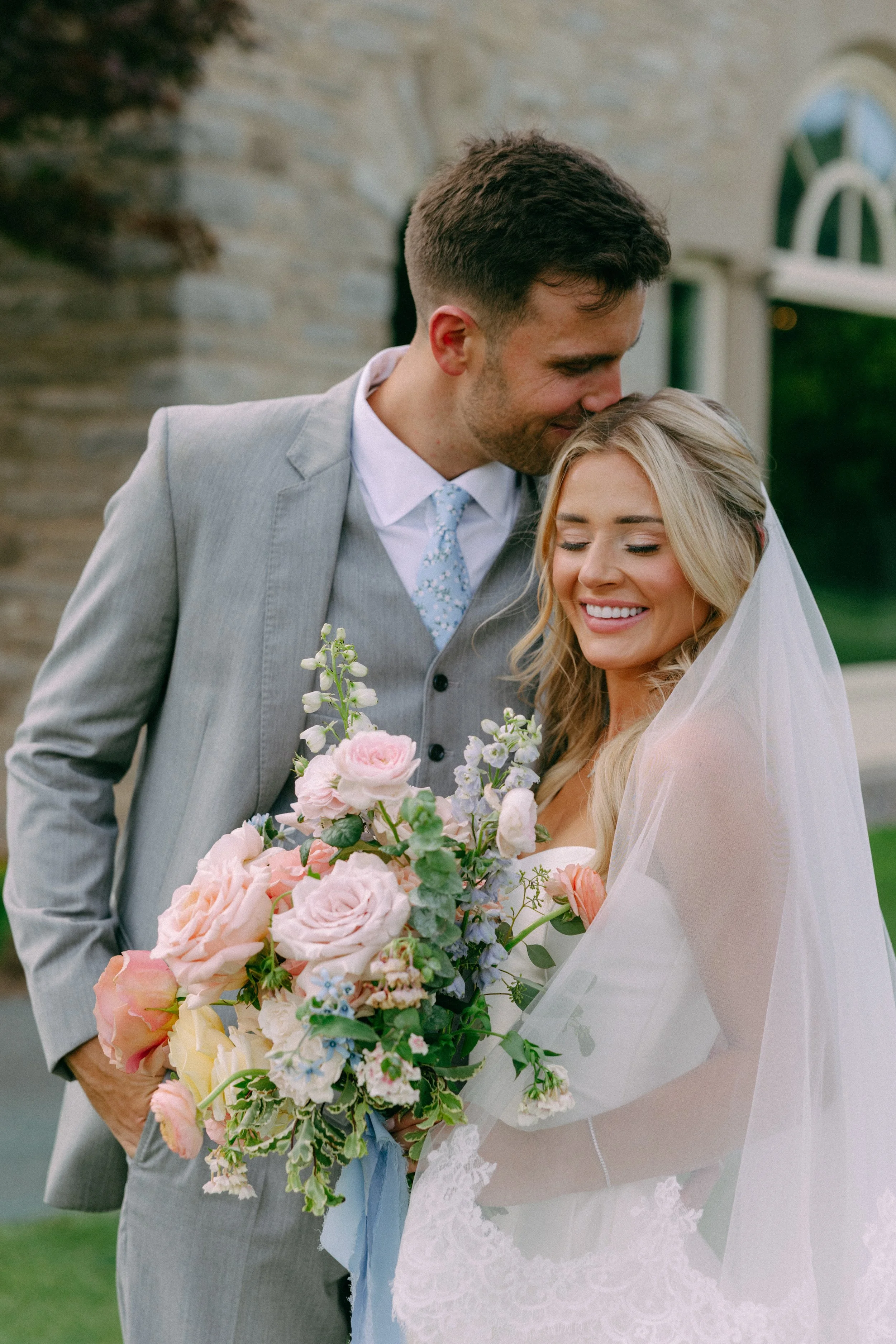 A newlywed couple with the groom leaning his forehead against the bride's head, both smiling, as she holds a large bouquet of pink and white flowers, outside near a stone building.