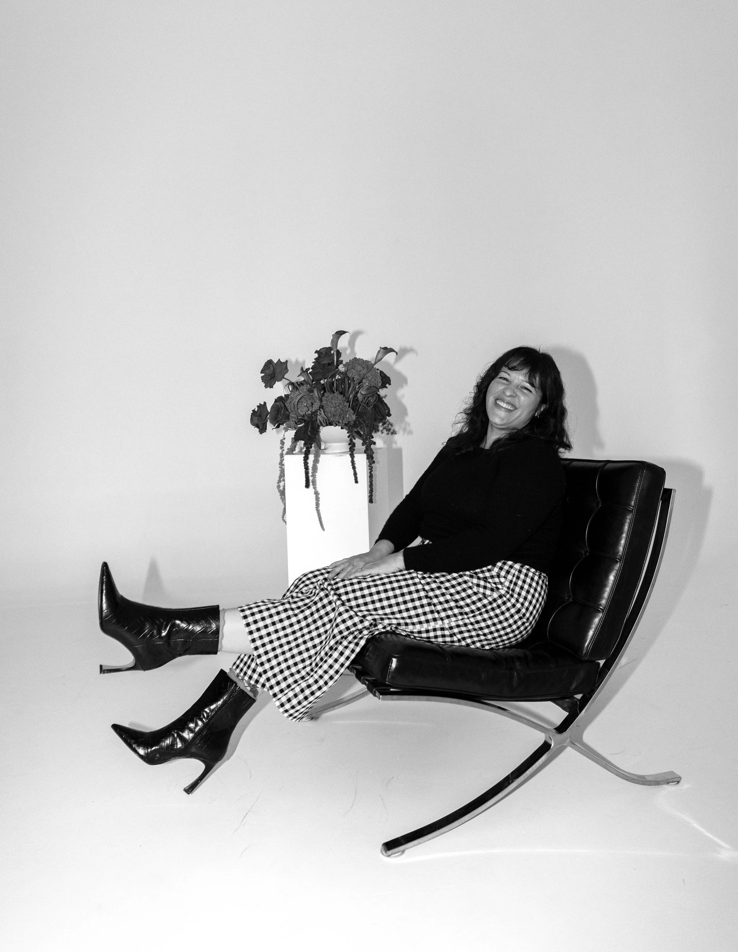 A woman sitting on a modern black leather chair, smiling, with a tall white vase of flowers beside her, in a minimalist setting.
