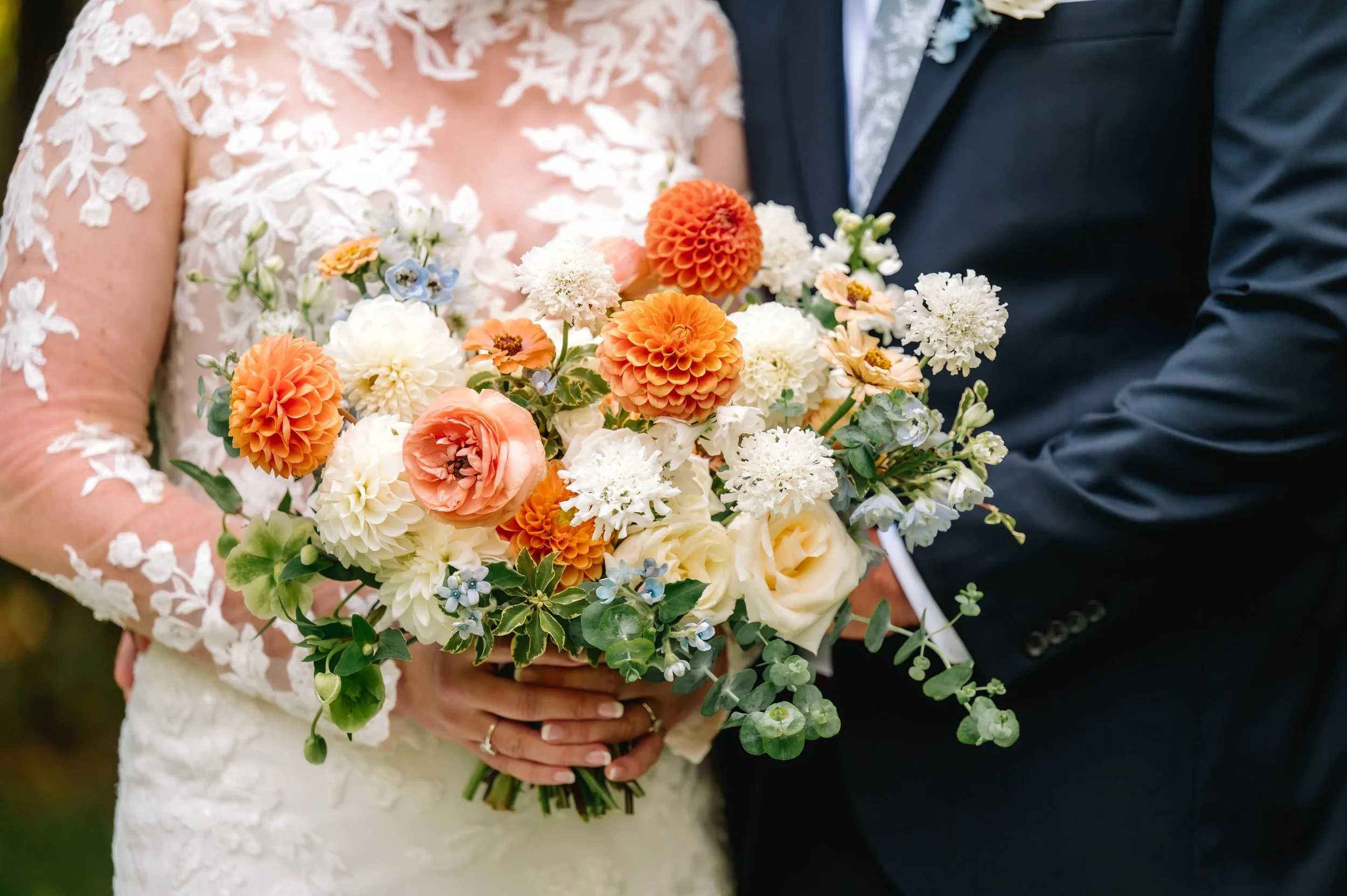 Close-up of a bride and groom holding a large, colorful bouquet of flowers during their wedding. The bride is wearing a lace wedding dress and the groom is dressed in a dark suit.