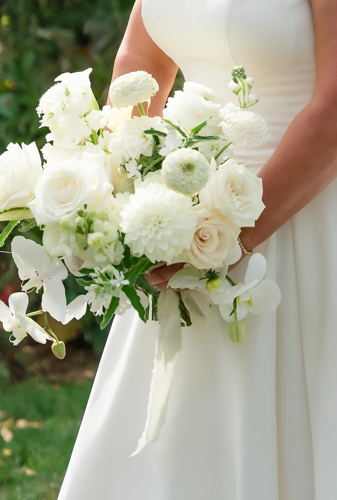 A bride in a white wedding dress holding a bouquet of white flowers, including roses and orchids, outdoors.