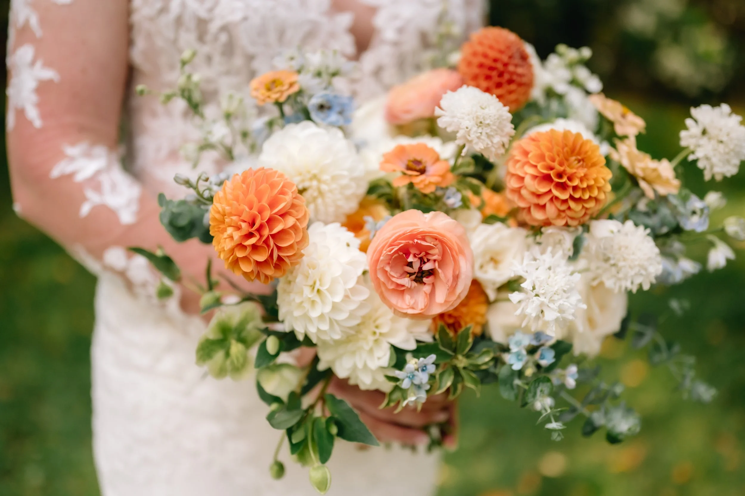 Person in a lace dress holding a colorful bouquet of orange, white, pink, and blue flowers outdoors.