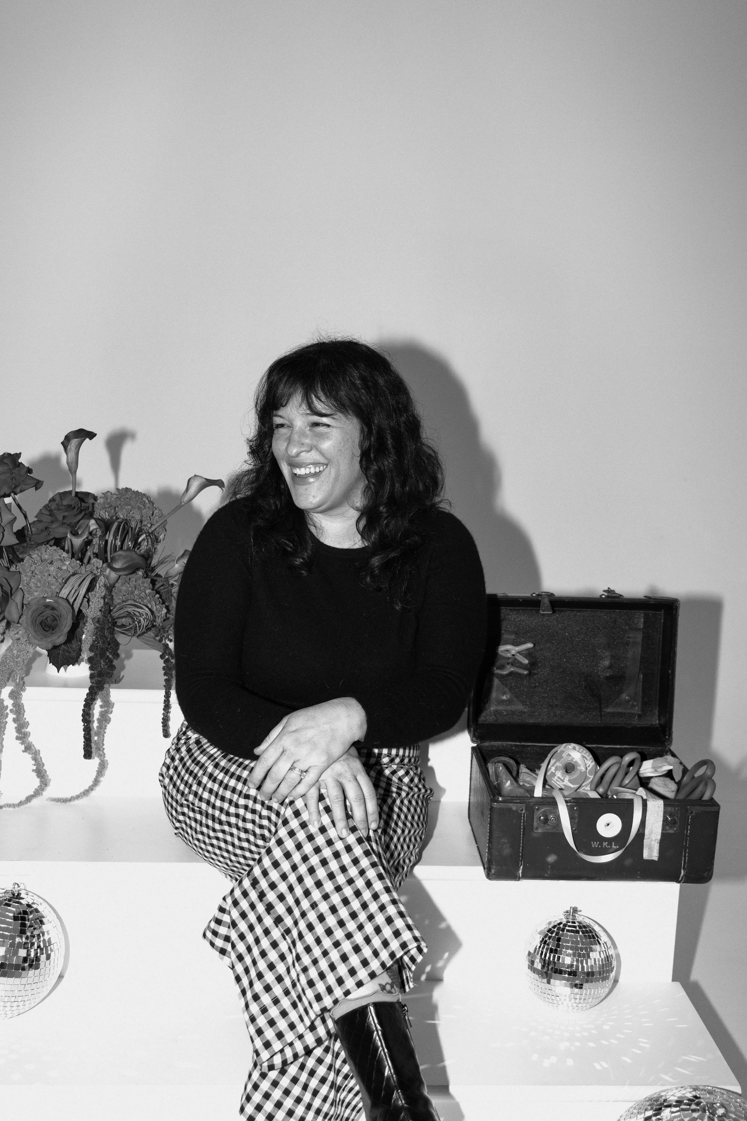 A woman with dark, wavy hair laughing, sitting barefoot with crossed legs, wearing a black top and checkered pants, next to a bouquet of flowers and a box of tools, with disco balls on the floor.