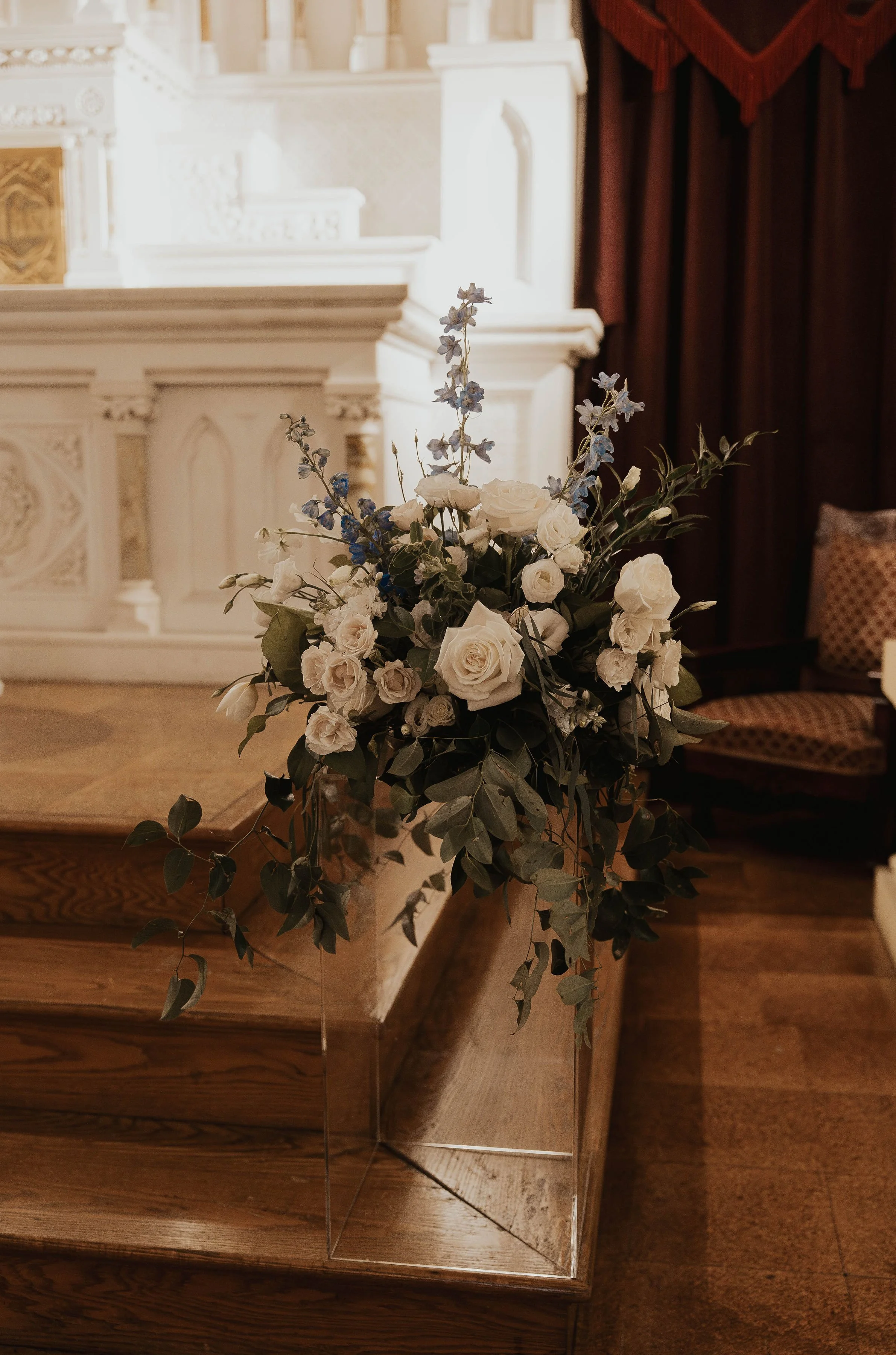 A floral arrangement with white roses, blue delphiniums, and greenery placed on a wooden platform in front of a white ornate altar in a church.