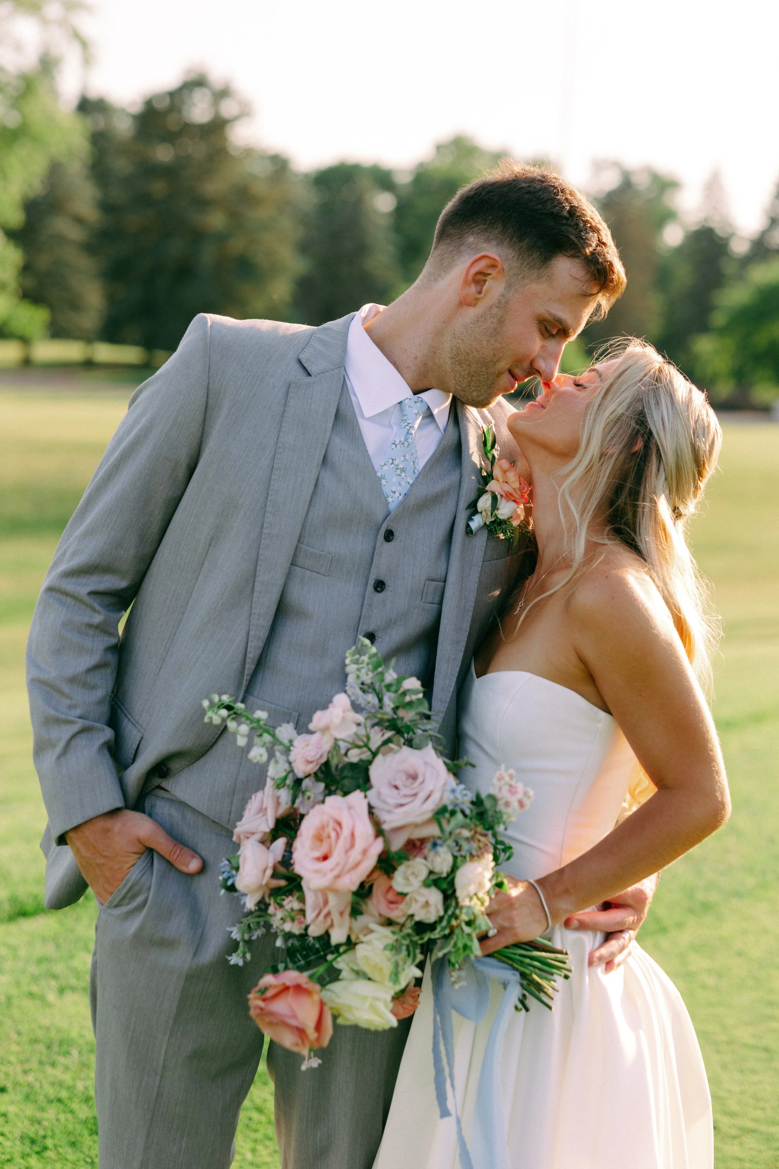 A newlywed couple on their wedding day, standing outdoors on a grassy field with trees in the background, gently leaning towards each other with their foreheads touching, holding a bouquet of pink and white flowers.