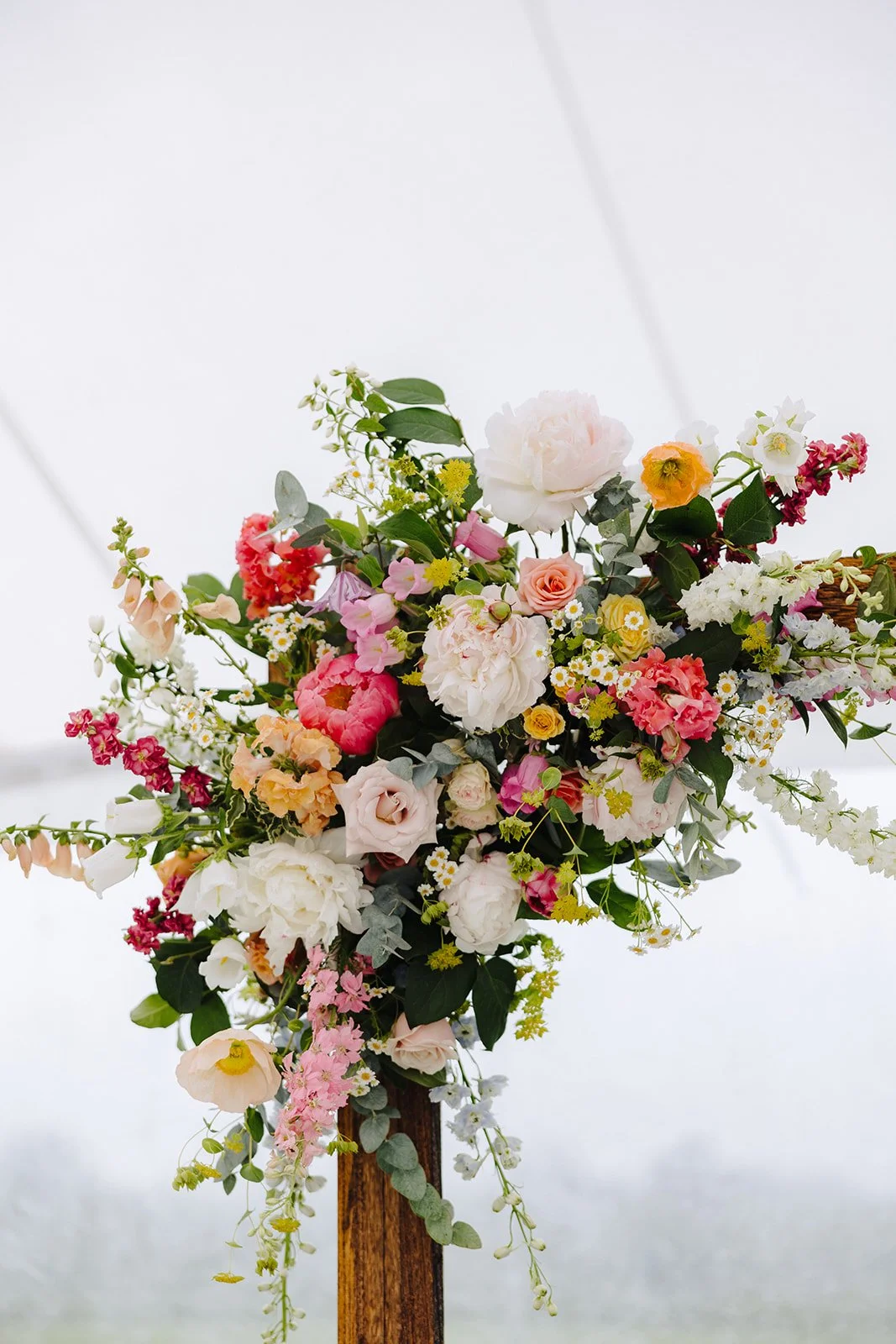 A bouquet of assorted colorful flowers including pink, white, yellow, peach, and red blossoms, arranged on a wooden stand against a bright, blurred background.