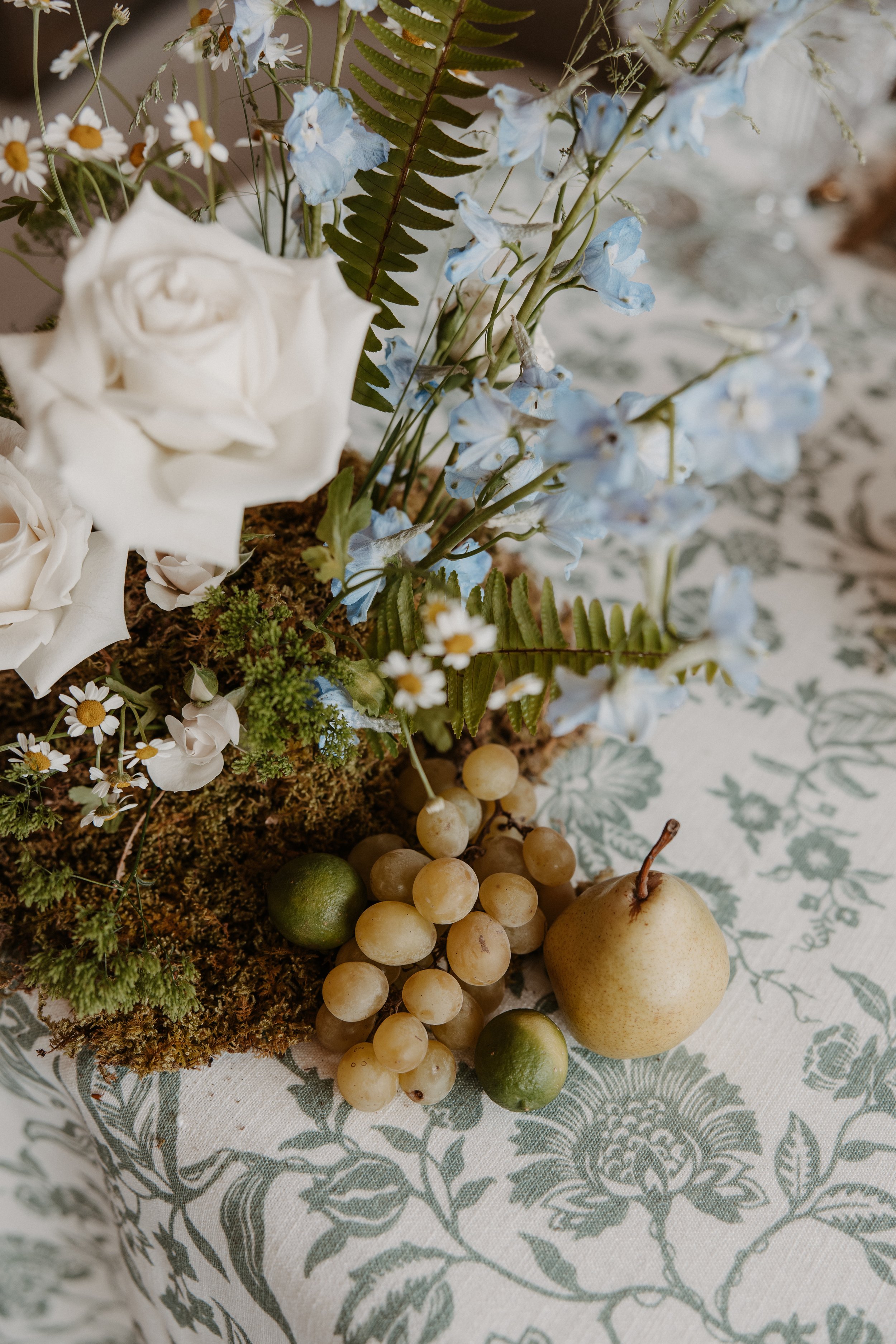 A floral arrangement with white roses, light blue flowers, small white daisies, and green ferns, placed on a decorative tablecloth with a floral pattern. There are also green and yellow pears, green limes, and a bunch of white grapes at the base of the arrangement.