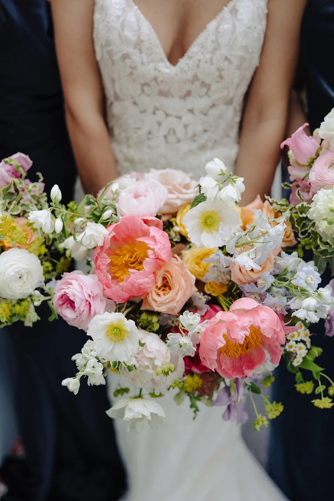 Close-up of a woman in a lace wedding dress holding a colorful bouquet of pink, white, peach, and yellow flowers.