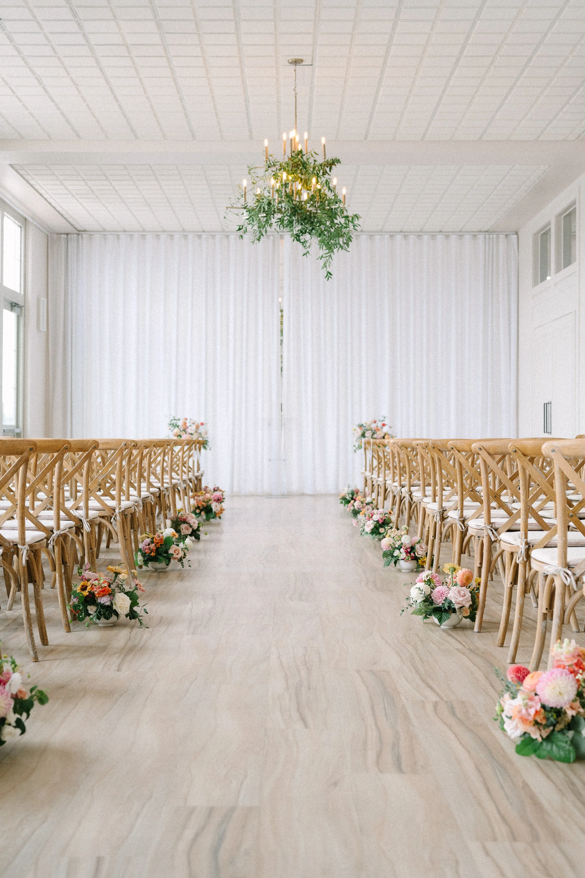 An elegant wedding ceremony setup with rows of wooden cross-back chairs decorated with floral arrangements along the aisle, a white curtain backdrop, and a chandelier with greenery hanging from the ceiling.