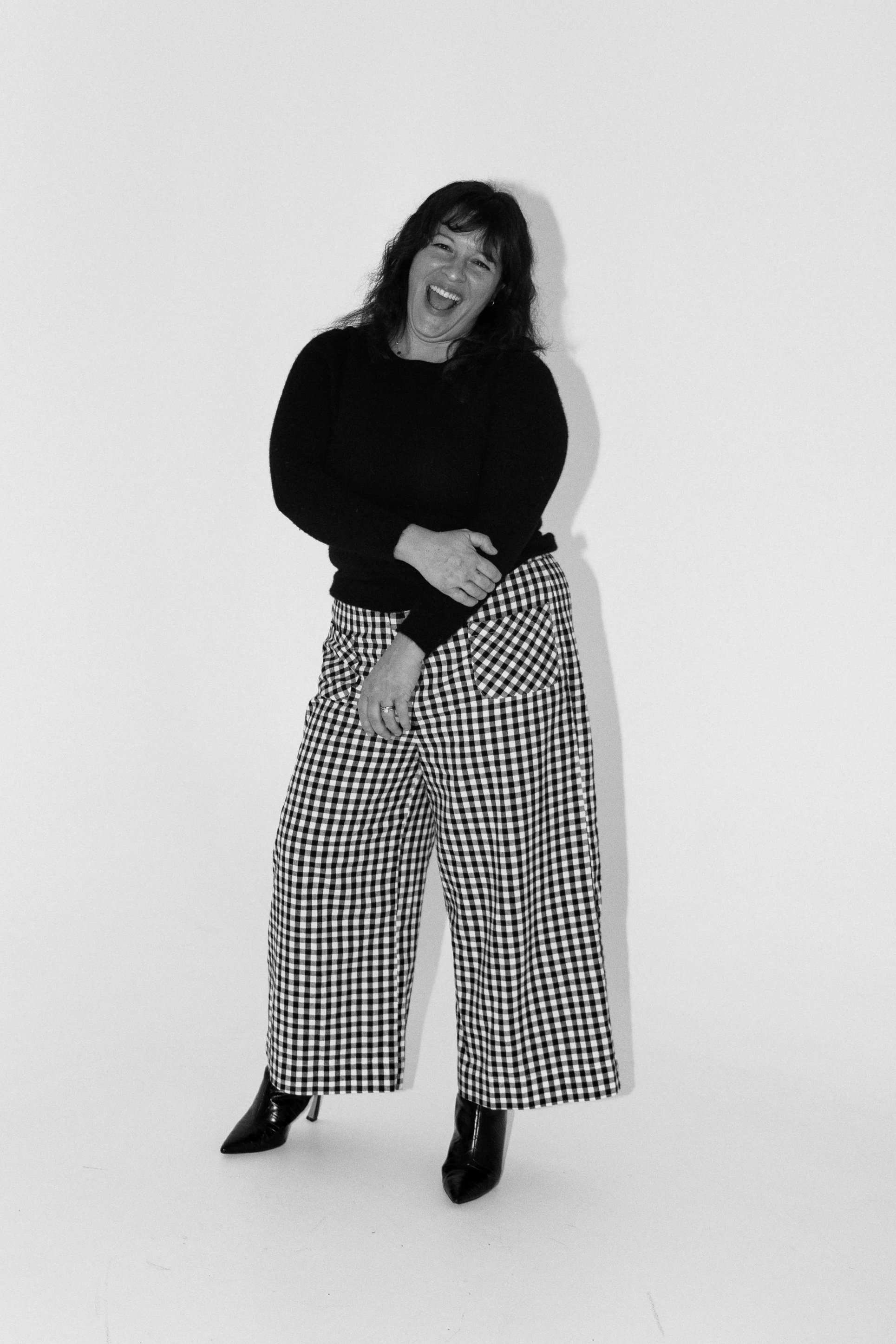 A woman smiling and laughing, wearing a black top, checkered wide-leg pants, and black ankle boots, standing against a plain white background.