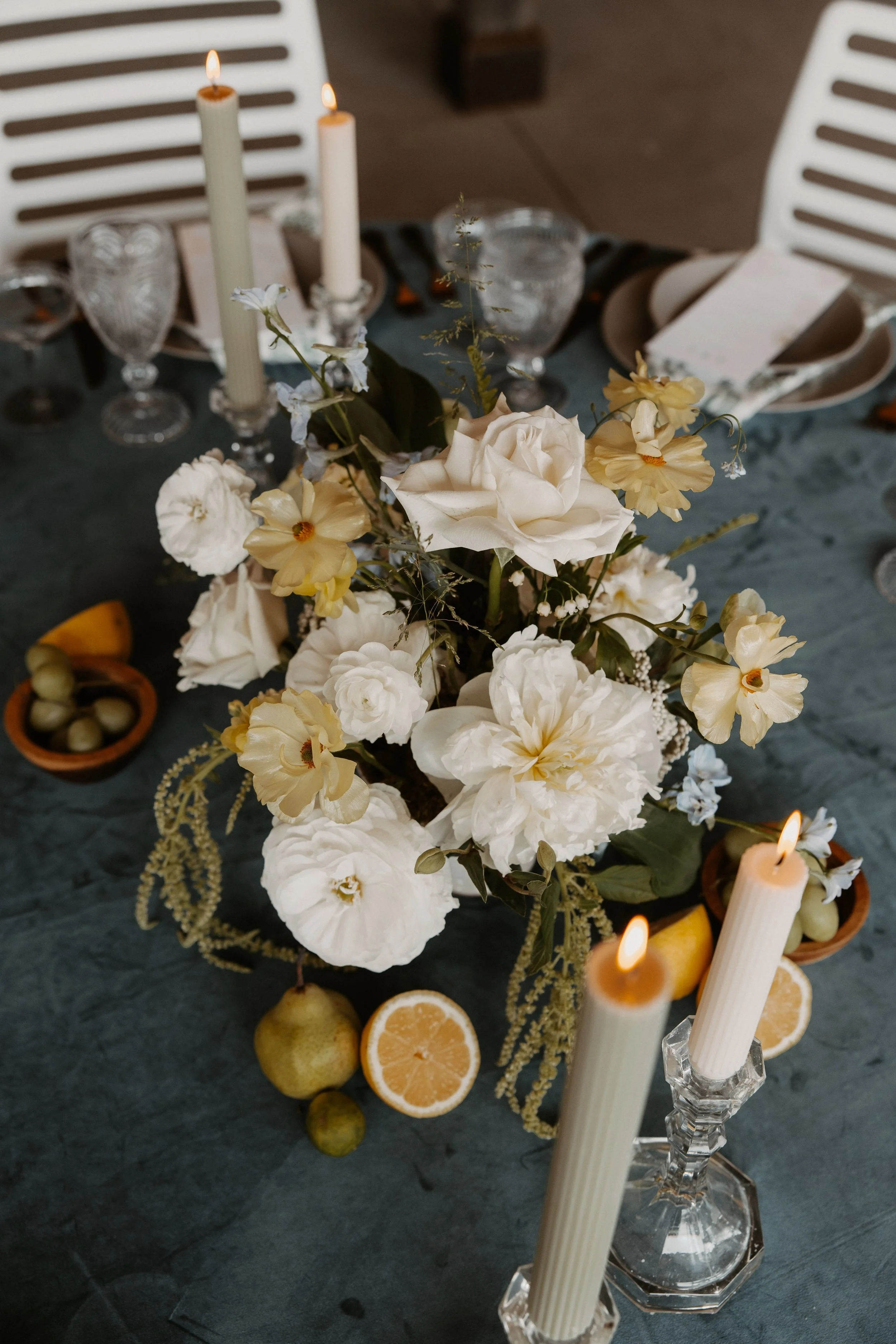 A table centerpiece with white and yellow flowers, surrounded by candles, lemons, and limes, set on a dark tablecloth with plates and glasses.