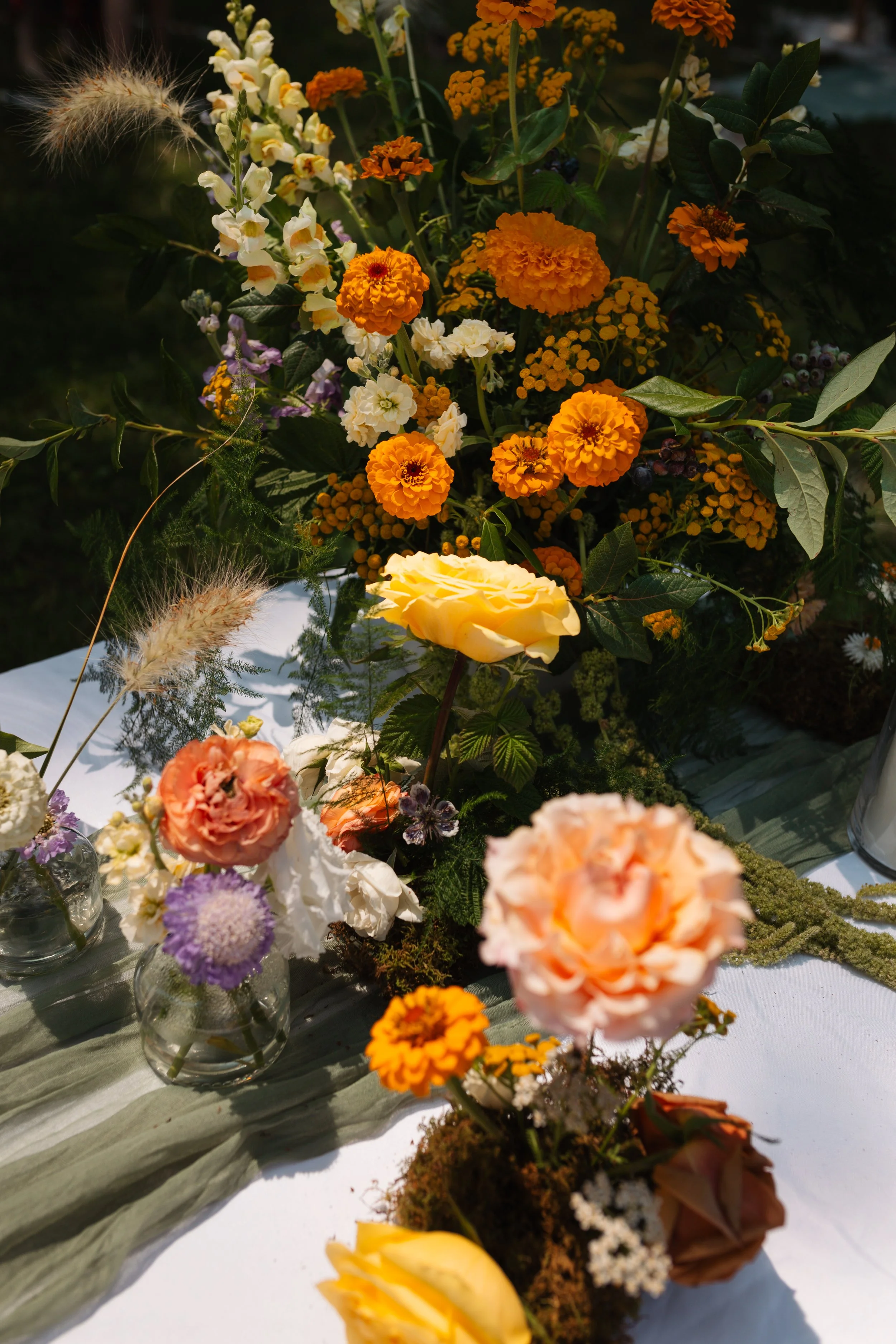 An arrangement of colorful flowers, including orange marigolds, yellow roses, white blossoms, and pink carnations, with greenery. Some flowers are in vases on a table covered with a white cloth.
