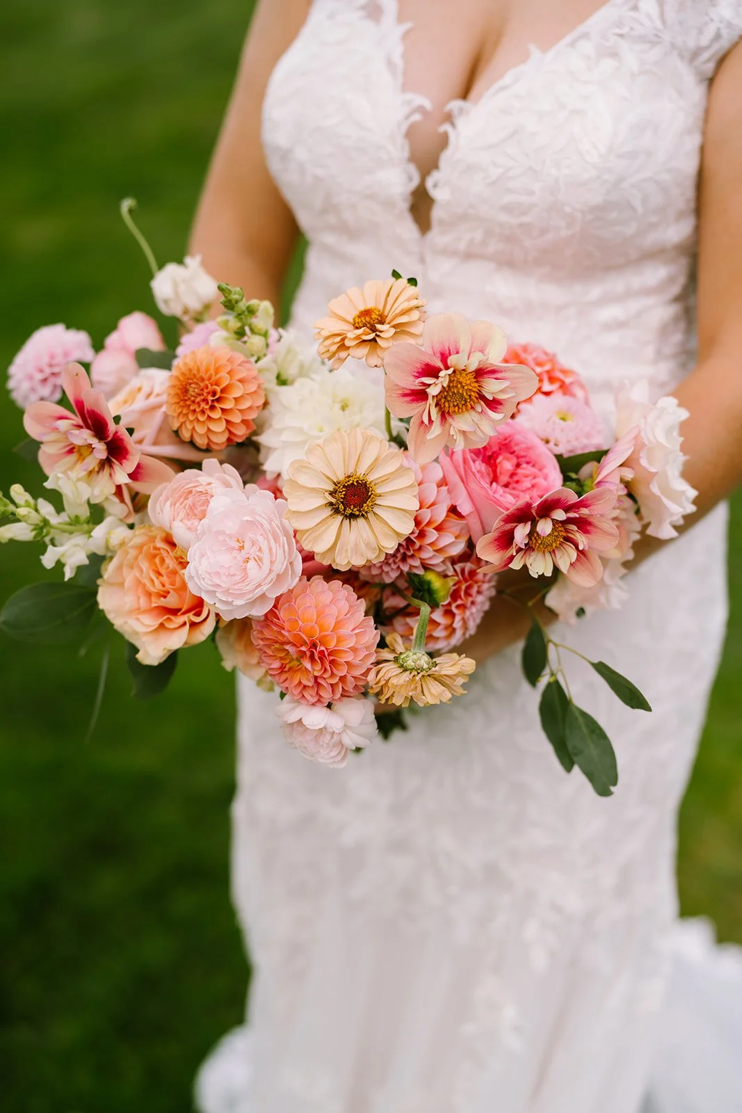 A bride holding a bouquet of pink and peach-colored flowers, including dahlias and zinnias, in front of her white lace wedding dress.