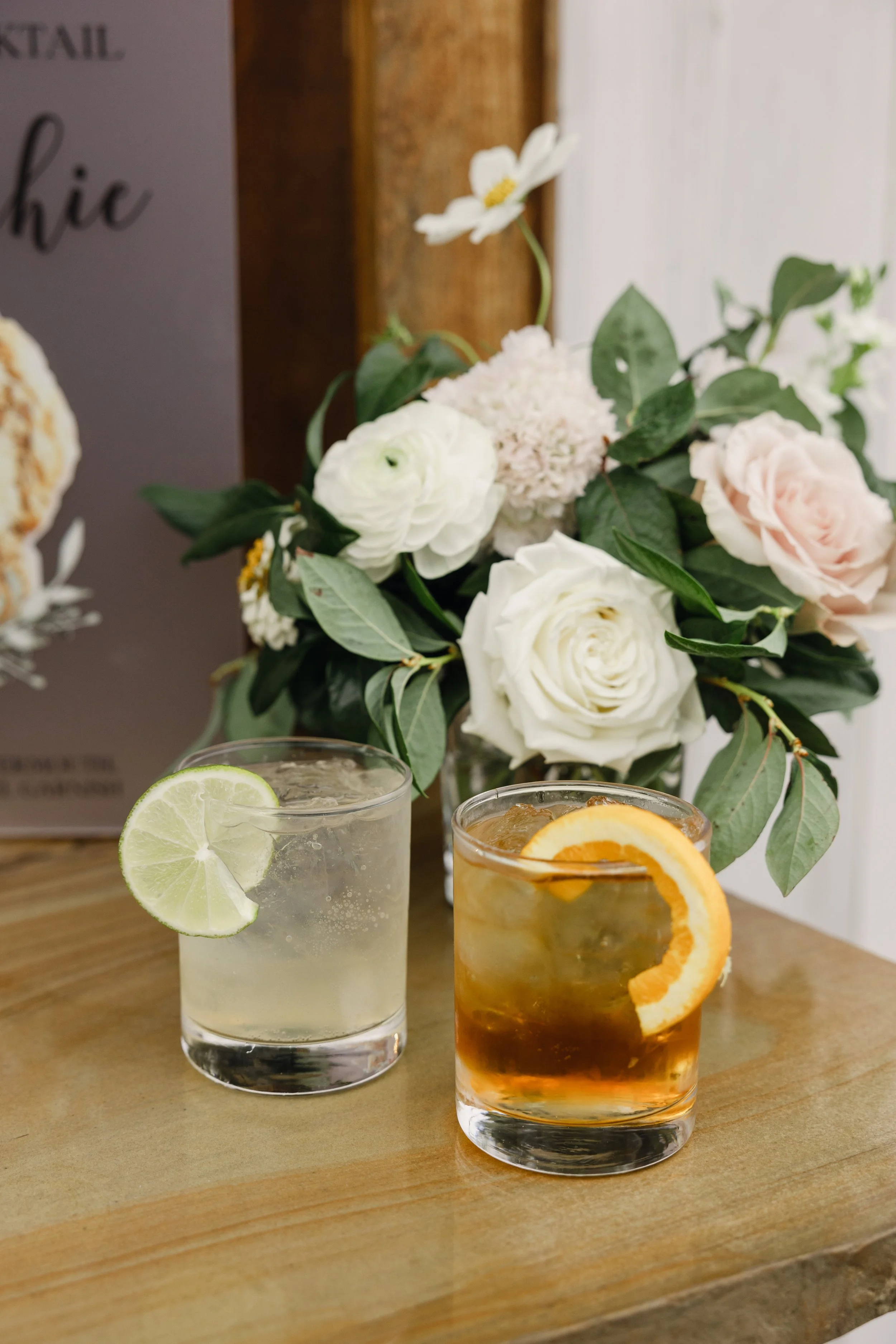 Two cocktail drinks with lemon and orange slices garnished, placed on a wooden surface with a bouquet of white and pink flowers in the background.