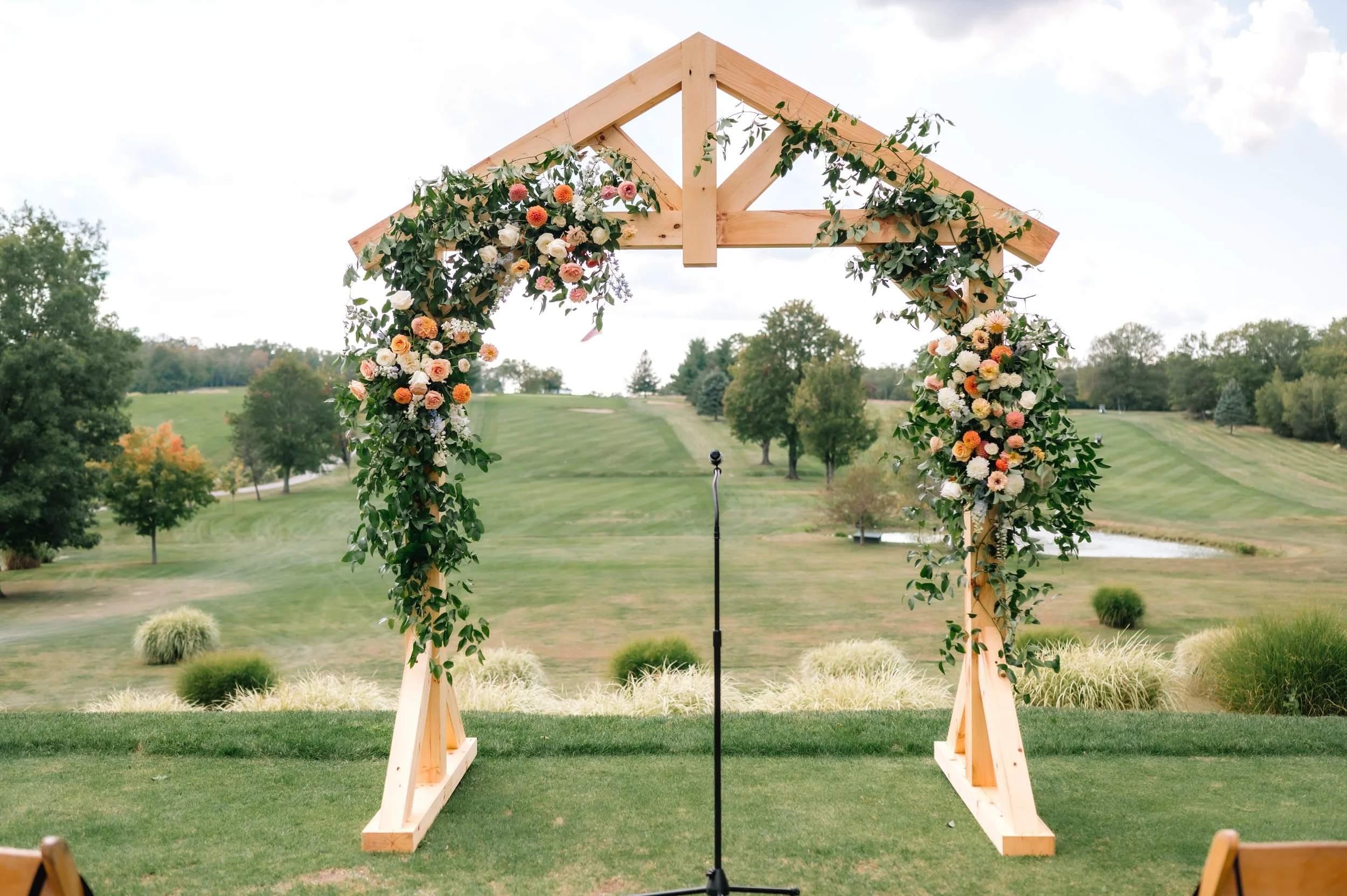 Wooden wedding arch decorated with flowers, set outdoors on a grassy hill with trees and a pond in the background.