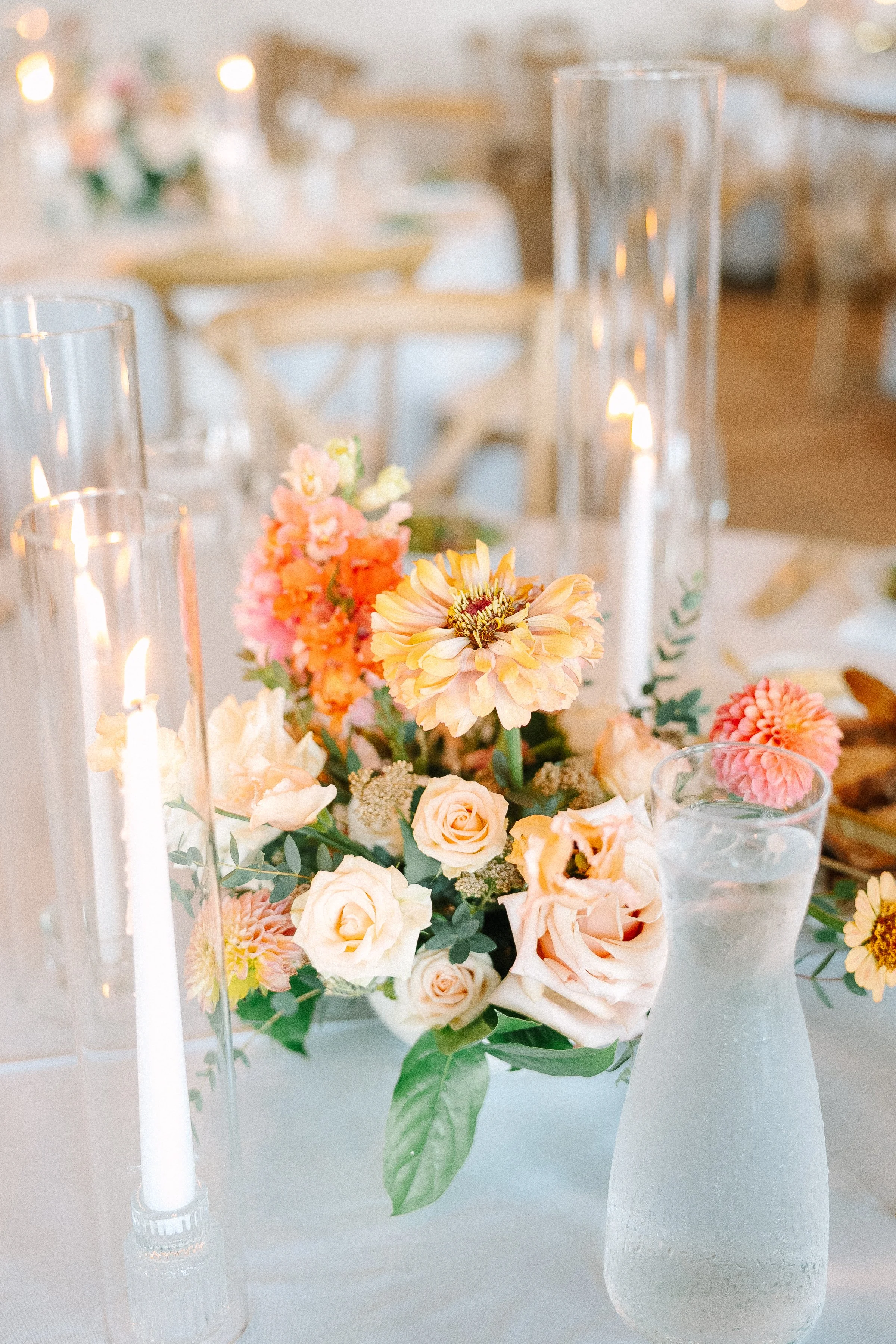 A floral centerpiece with pink, peach, and cream flowers on a white table, surrounded by tall glass candle holders and a frosted glass water pitcher, in a softly lit event setting.