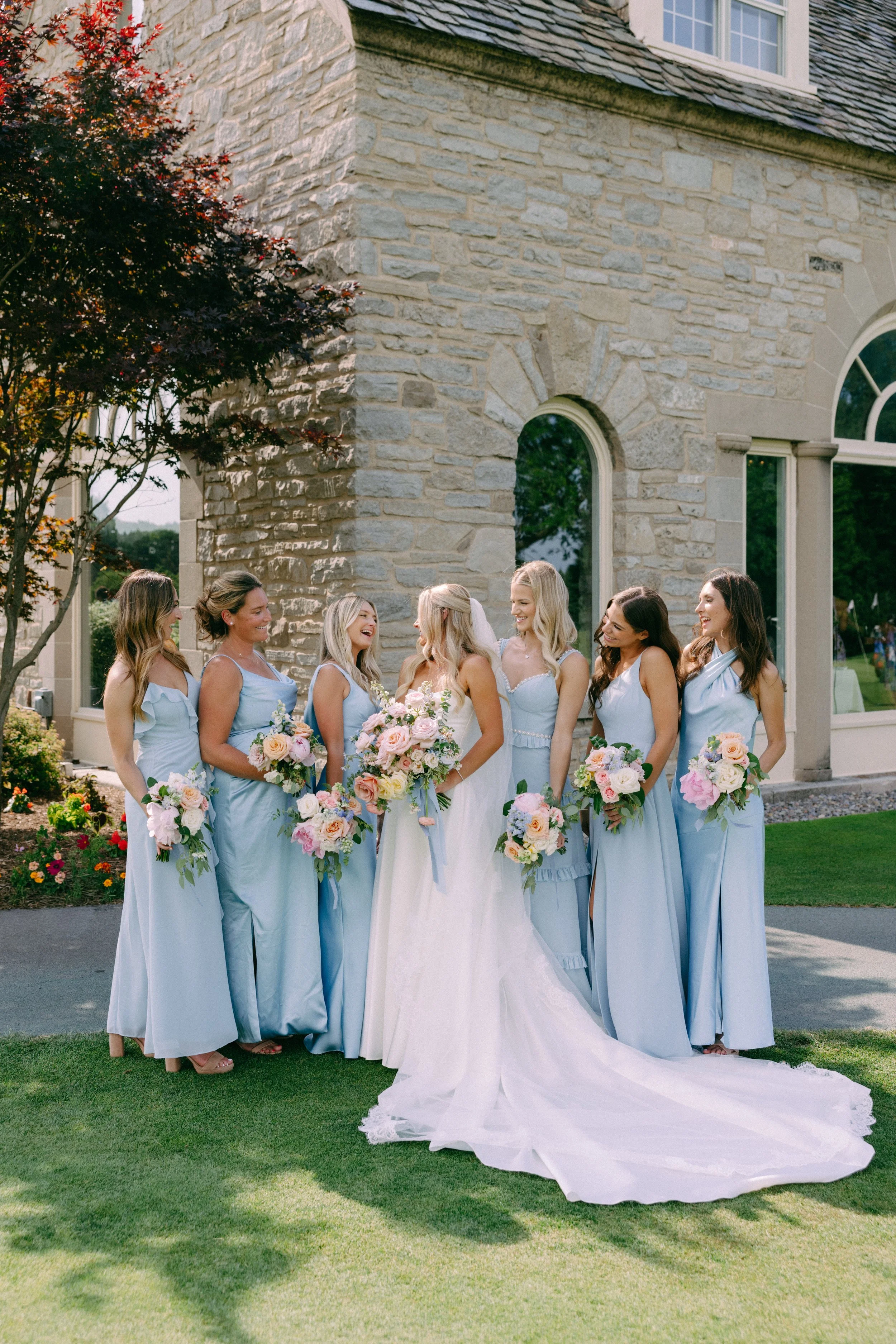 A bride in a white wedding dress talking to her bridesmaids dressed in light blue dresses, outdoors in front of a stone building, holding bouquets of pink and white flowers.