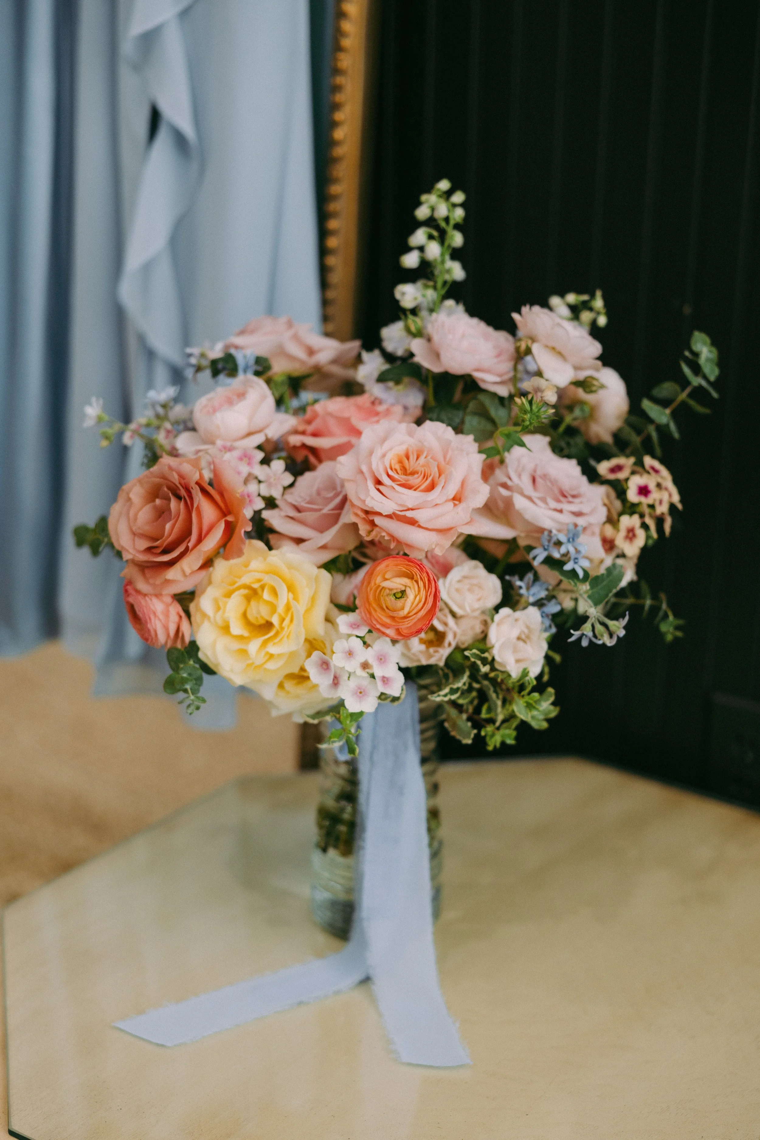 A bouquet of pink, yellow, and peach roses with some blue and white flowers in a glass jar, decorated with a light blue ribbon, placed on a beige surface.