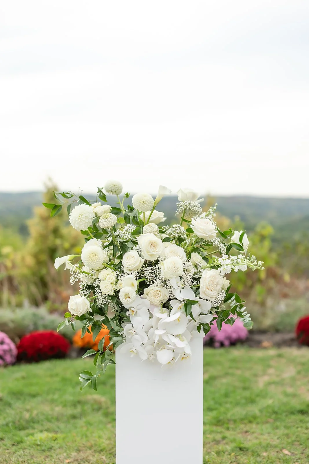 White floral arrangement with roses, lilies, and baby's breath on a pedestal outdoors, with a blurred garden background.