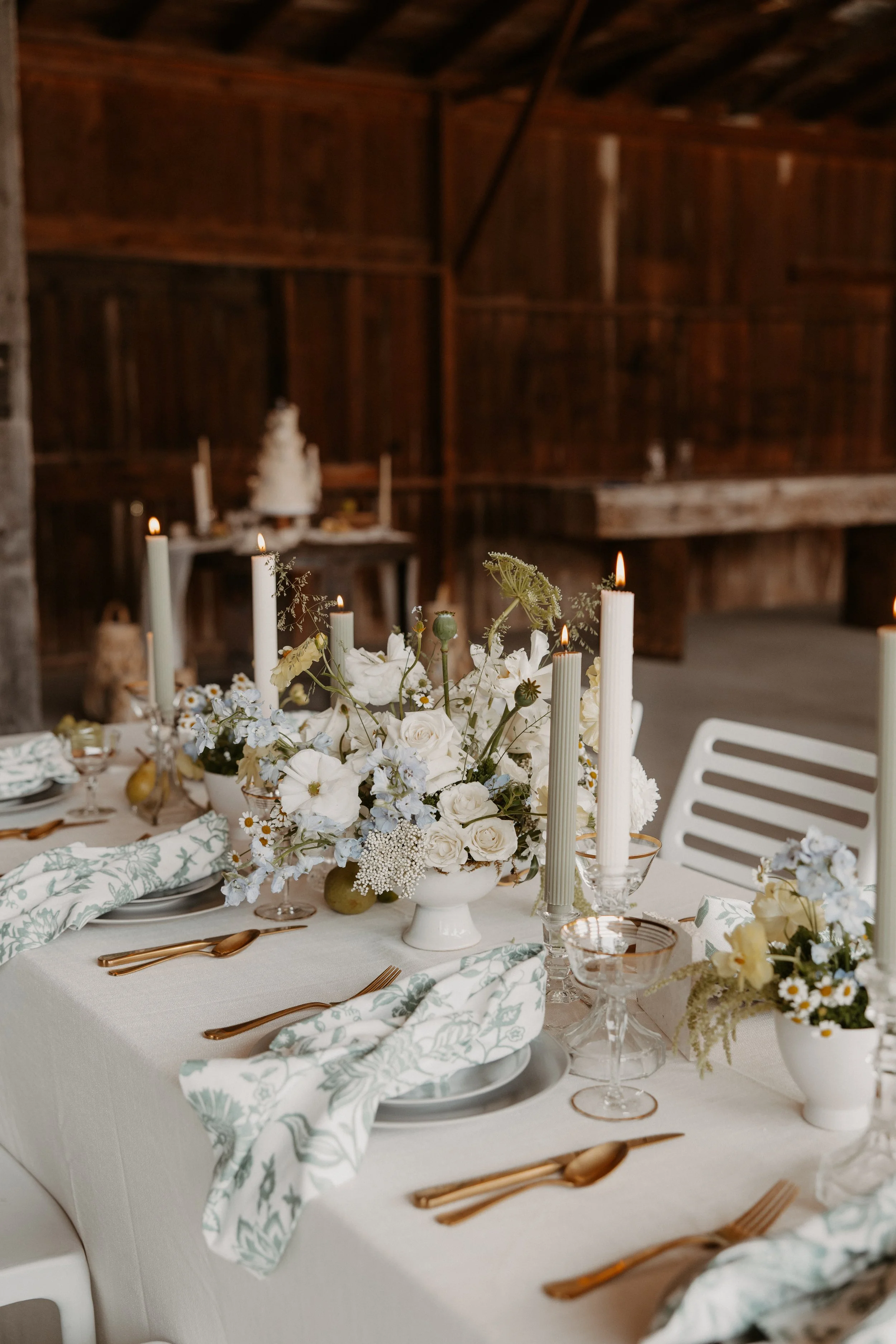 Decorated table with white and pale green floral centerpiece, candles, gold flatware, and patterned cloth napkins in a rustic wooden barn setting.