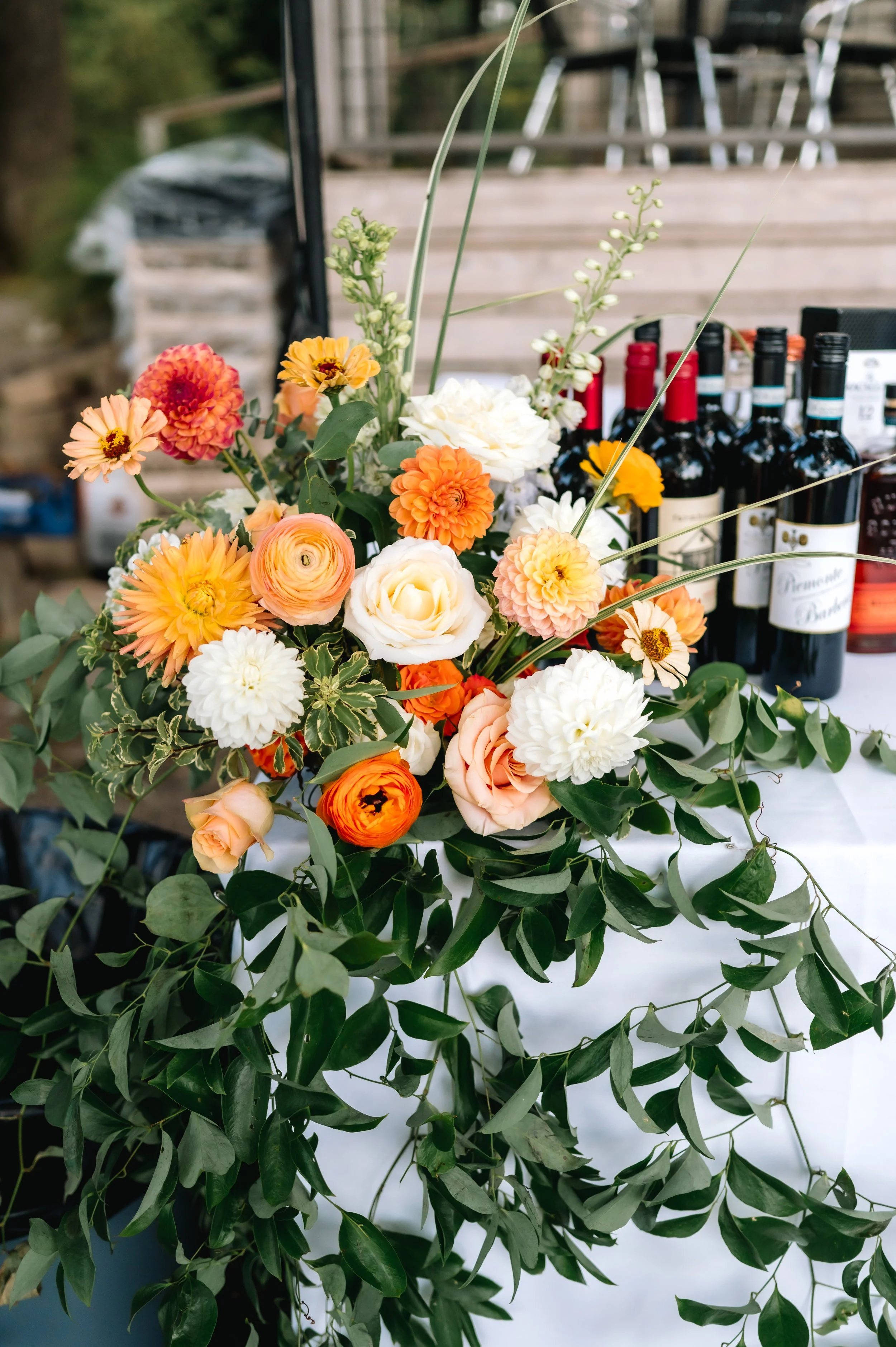 A colorful bouquet of flowers including orange, white, and peach blossoms, with greenery, set on a table with bottles of wine in the background.
