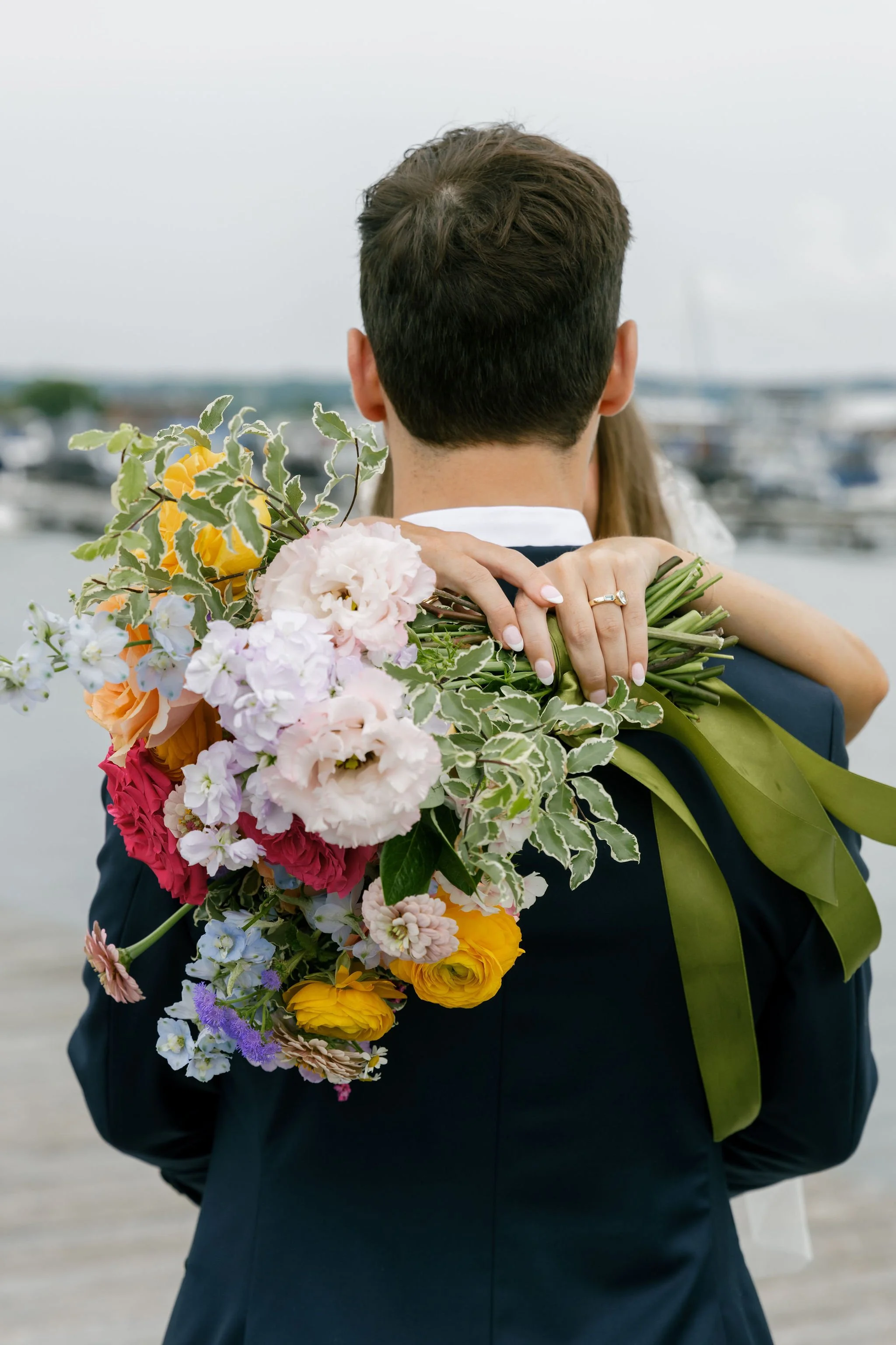 A groom in a navy suit holding a bride in his arms, with her arms wrapped around his neck, at a marina. The bride's hand, with a wedding ring, rests on his shoulder, holding a large bouquet of colorful flowers.