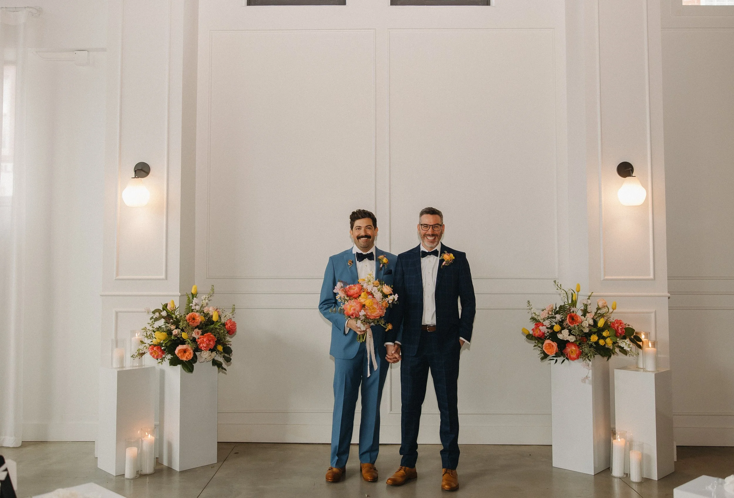 Two men in suits holding hands and a bouquet of flowers, standing in an elegant, brightly lit room with white walls, floral arrangements, and candles.