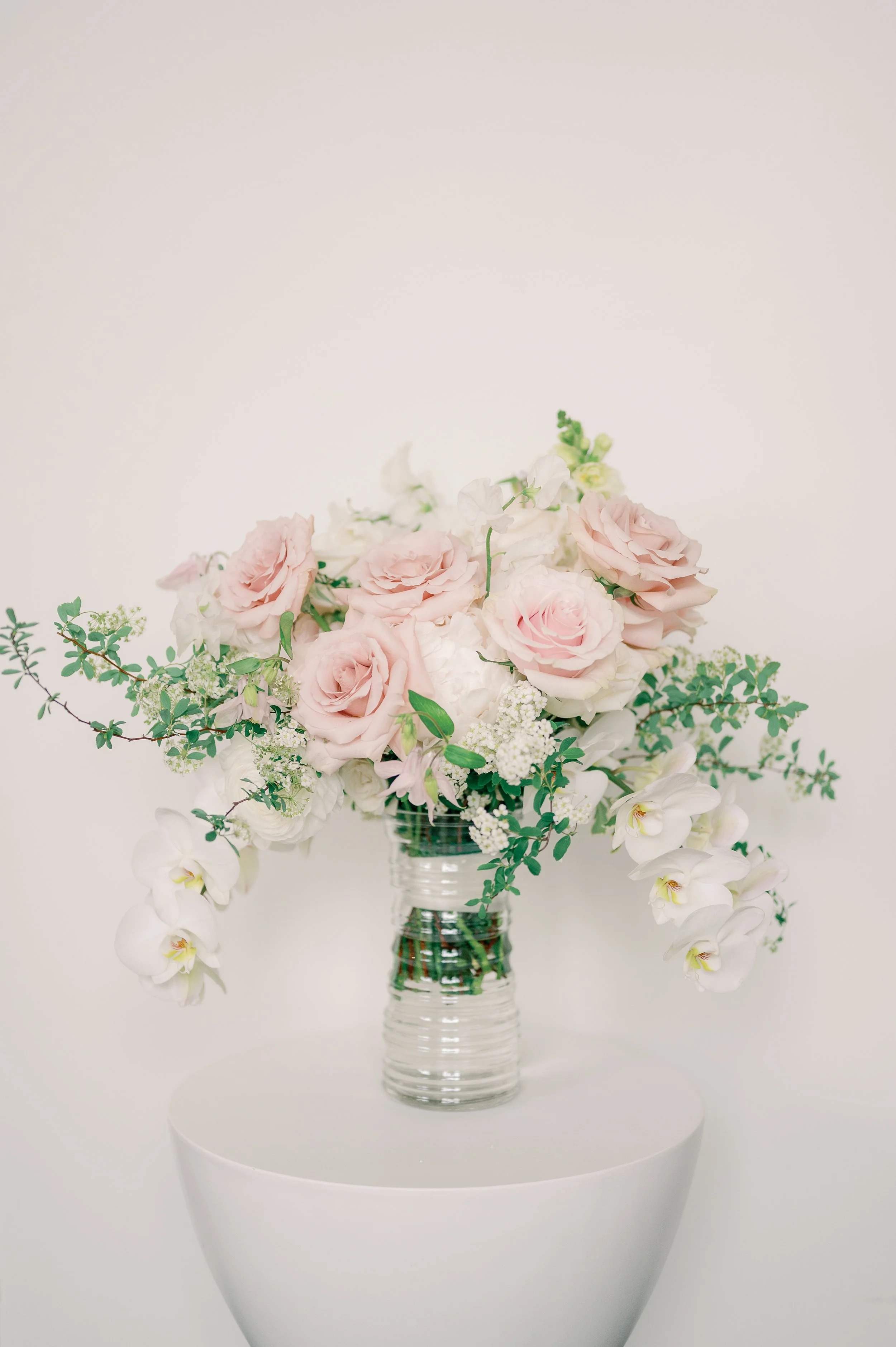 A floral arrangement with pink roses, white orchids, and other white and green flowers in a clear glass vase on a white surface against a white background.