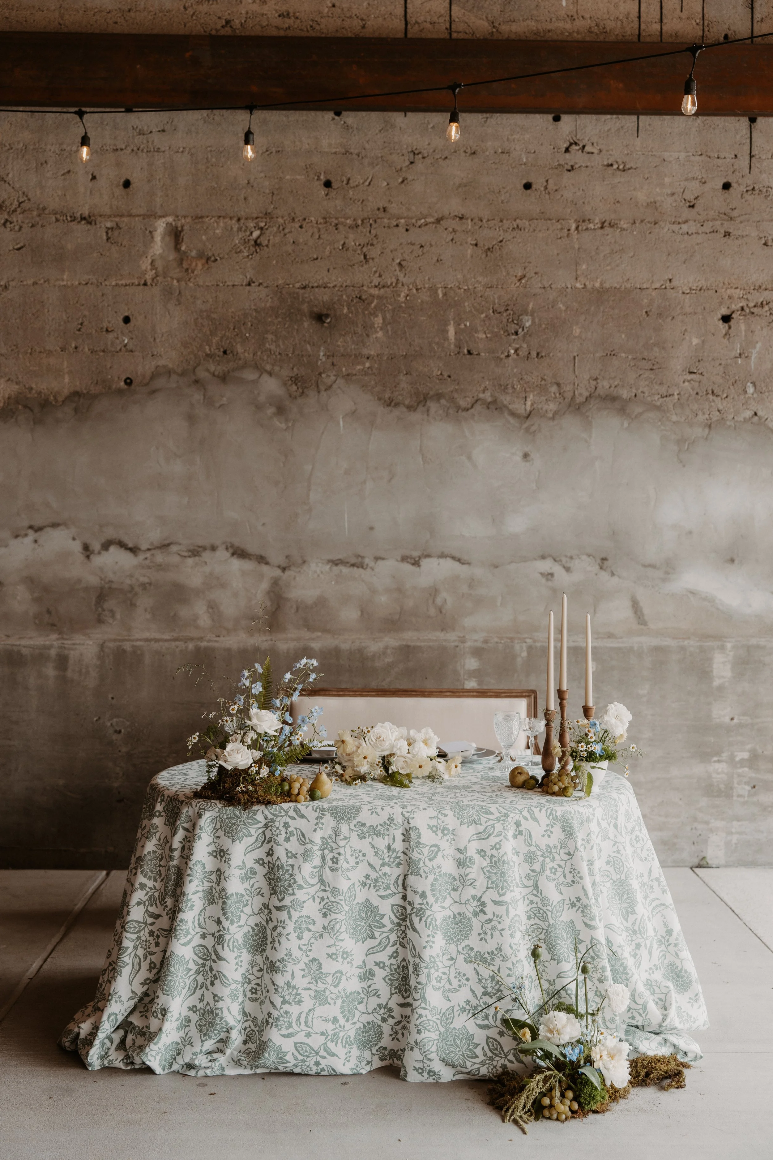 A decorated round table with a floral tablecloth, white and pale blue flowers, tall candles, and glassware, set against a textured gray wall with string lights overhead.