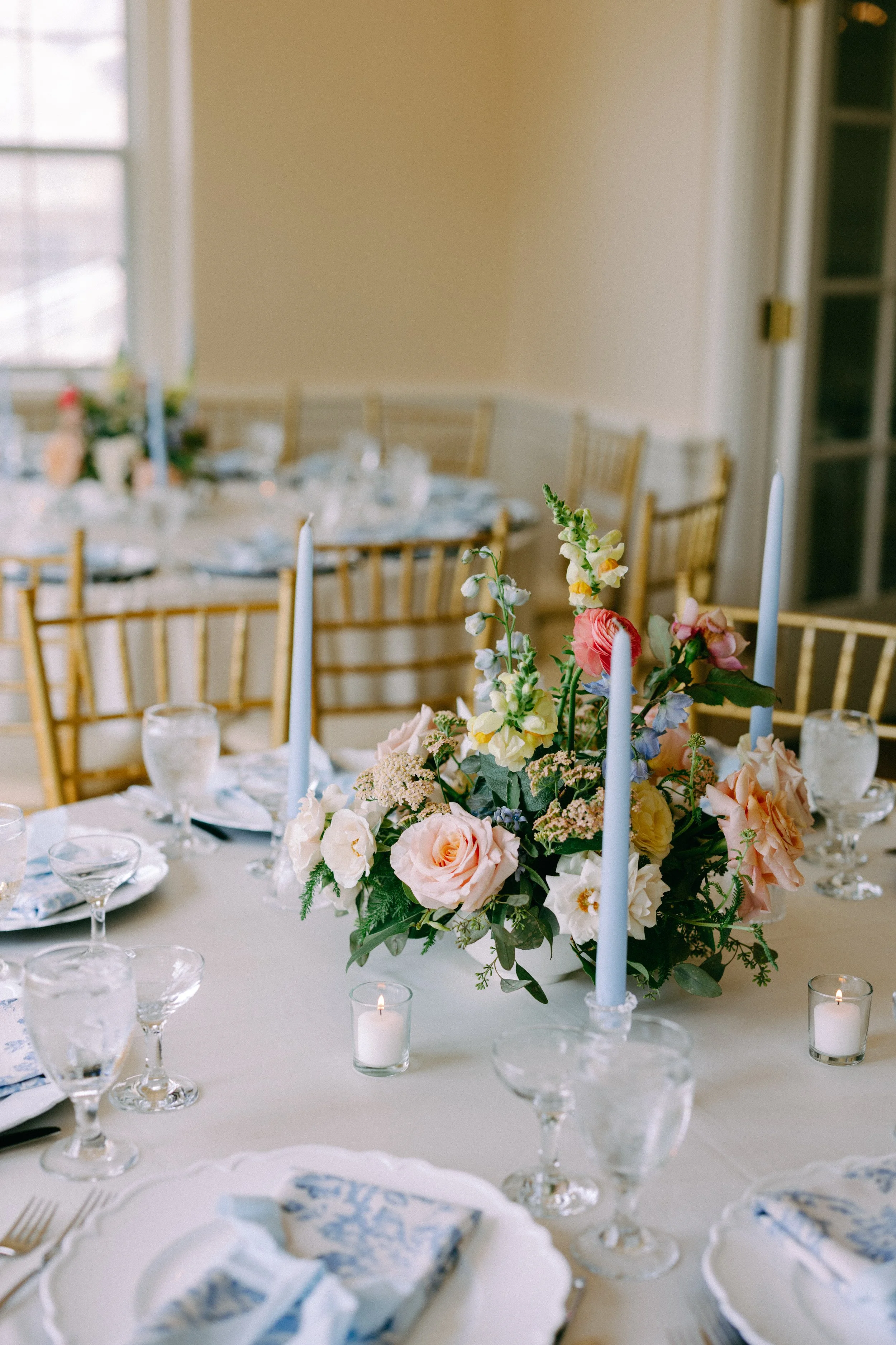 Elegant dining table with a floral centerpiece, blue taper candles, glassware, and table settings in a well-lit room.
