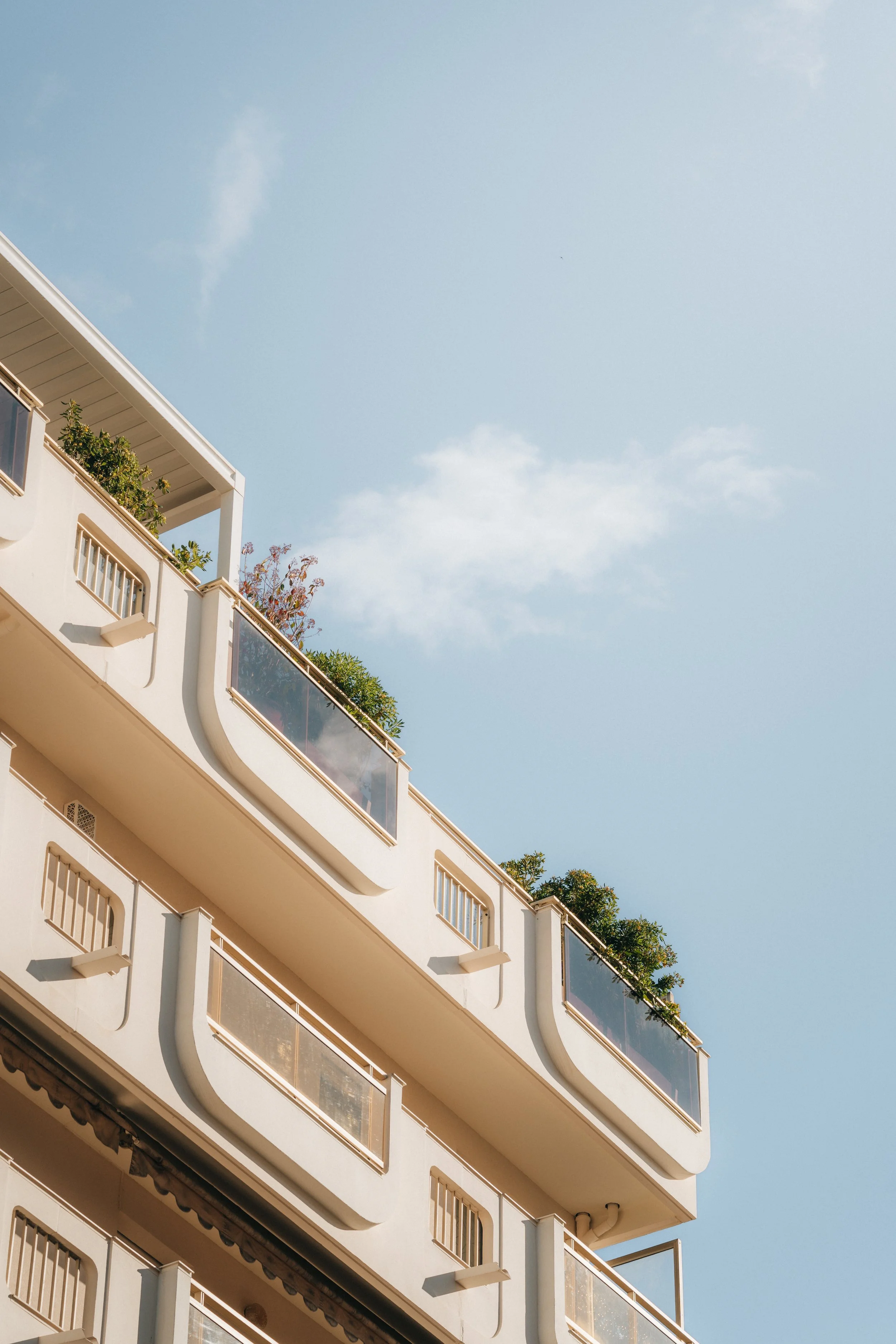 Vue d'un bâtiment blanc avec balcons en verre, vue partielle de plusieurs étages, sous un ciel bleu avec peu de nuages.