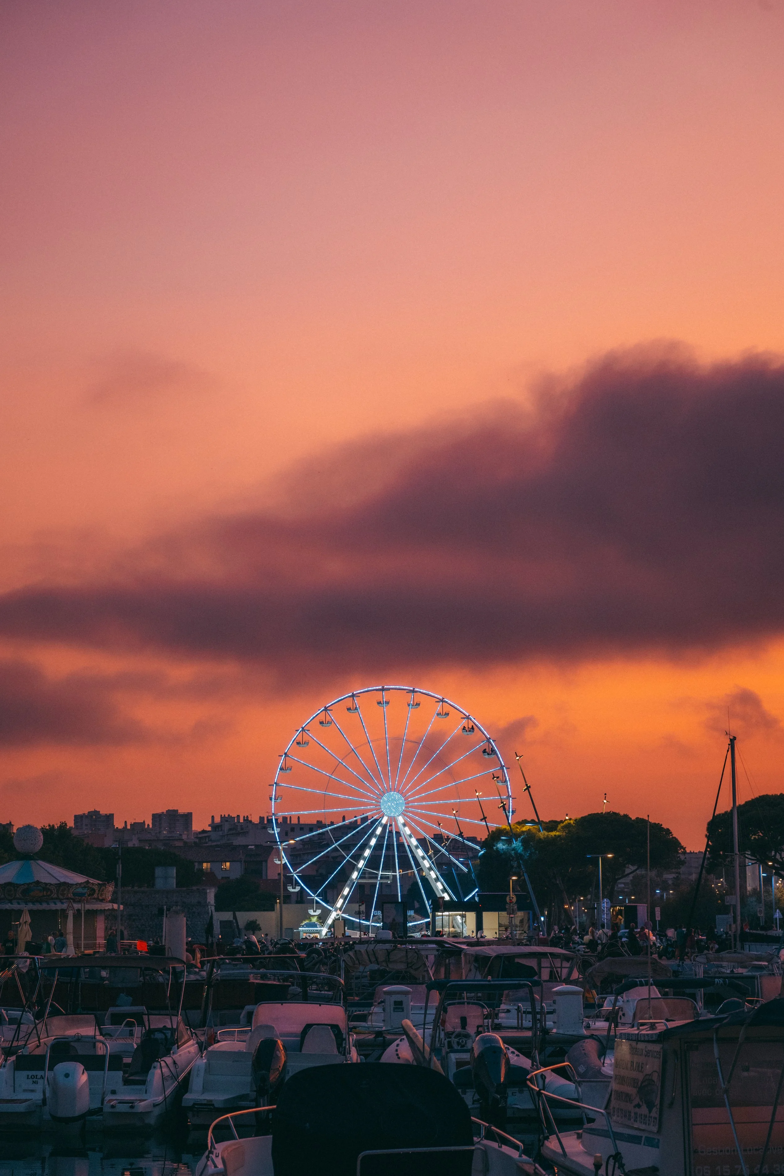 La photo montre une grande roue illuminée au parc d'attractions avec un ciel coloré de coucher de soleil, des bateaux au premier plan et une silhouette de ville en arrière-plan.